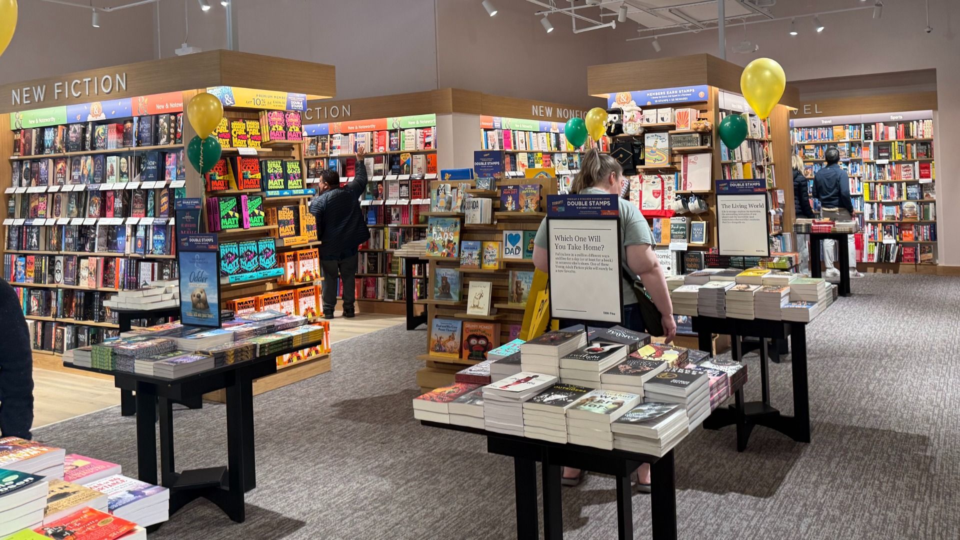 Inside a bookstore, the large "New Fiction" section features tall wooden shelves, colorful novels, display tables of books, and shoppers browsing among balloons.