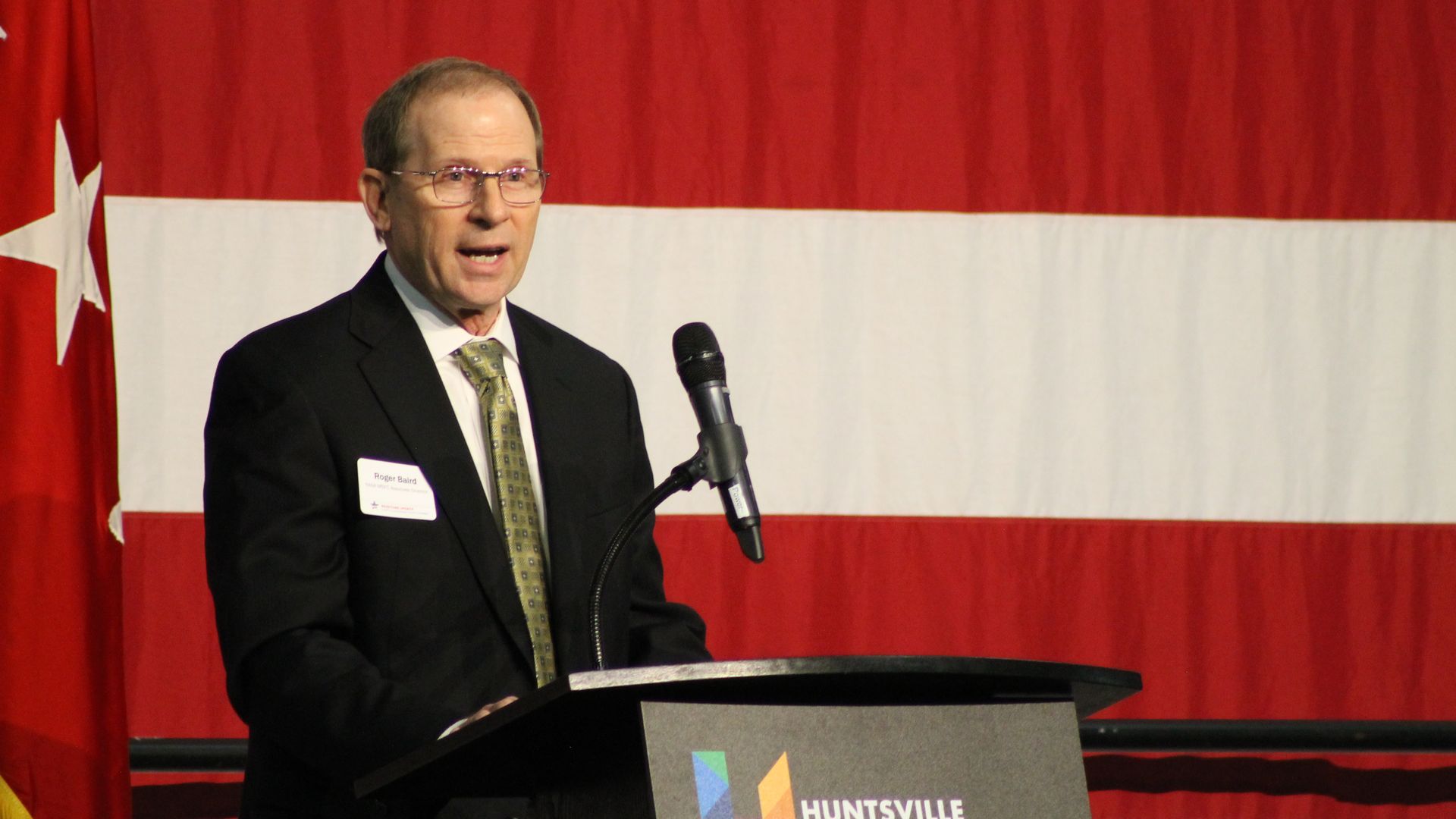 Man in black suit and green tie speaking at a podium with a microphone, standing in front of a large red and white striped flag with a white star. The podium sign reads "Huntsville Madison County".