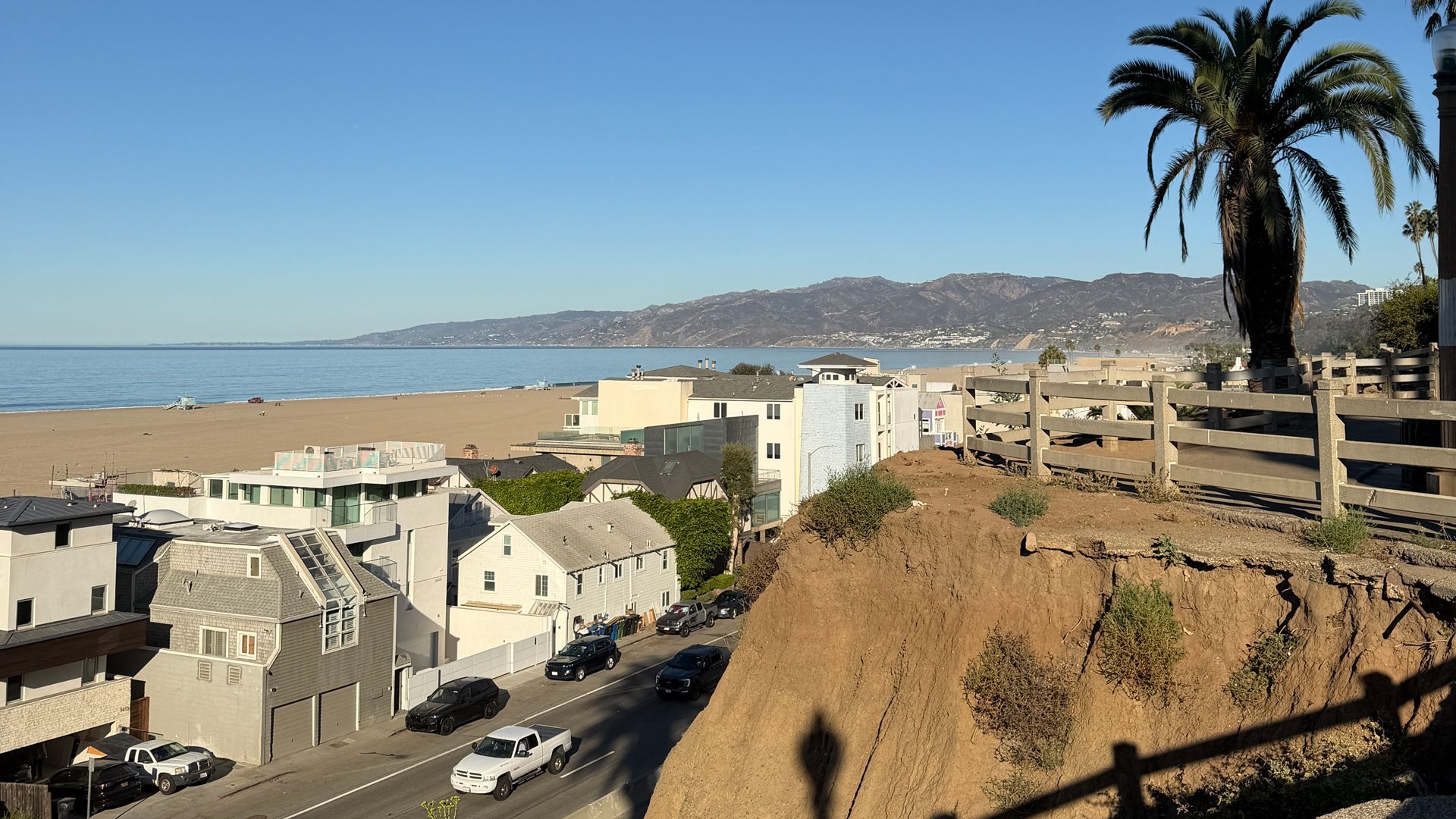 Sunny coastal scene with a sandy beach and blue sea on the left, row houses and a street with cars in the foreground, a wooden fence and a tall palm on the right, and distant mountains.