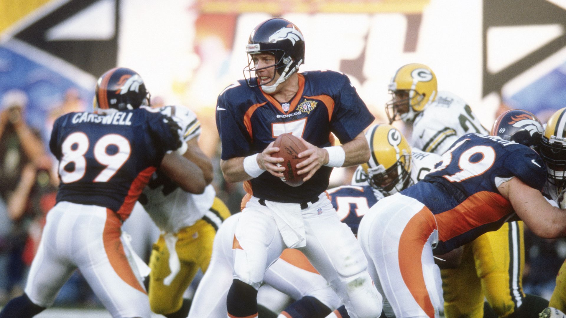 A man in a navy helmet holds on to a football while preparing to make a move on a field surrounded by other players. 