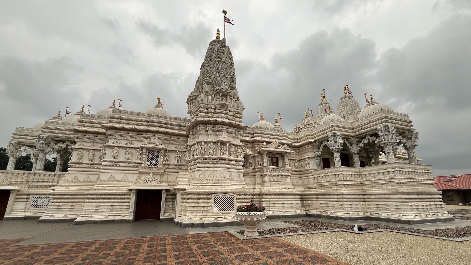 White temple with a tall central spire and accompanying domes, flags on top; highly detailed carvings on the façade, and a red-green tiled foreground under a cloudy sky.
