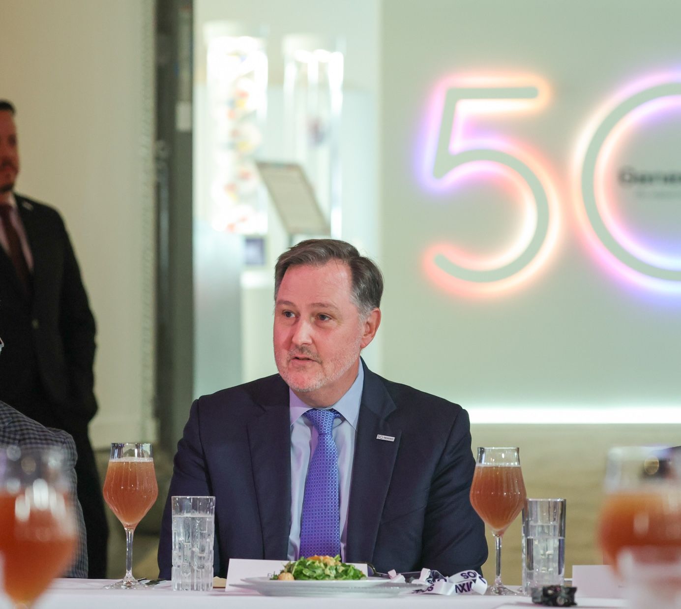 A man in a navy suit and tie sits at a banquet table with a plate of salad, glasses of orange beverage, and a neon 50 sign in the background.
