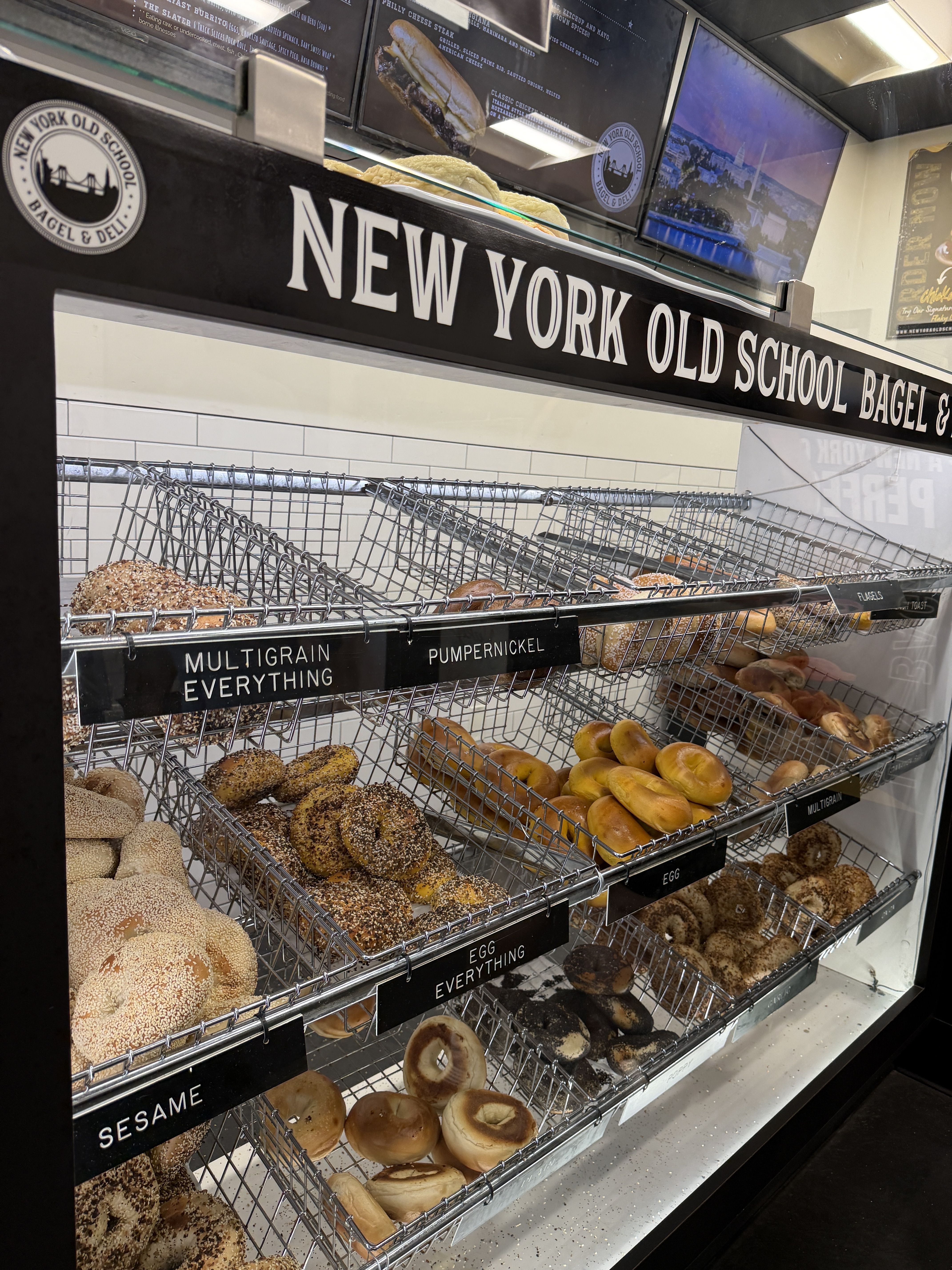 Bakery display at New York Old School Bagel & Deli: metal racks of bagels in baskets, including sesame, multigrain, and everything; large black sign and round logo above show the shop name.