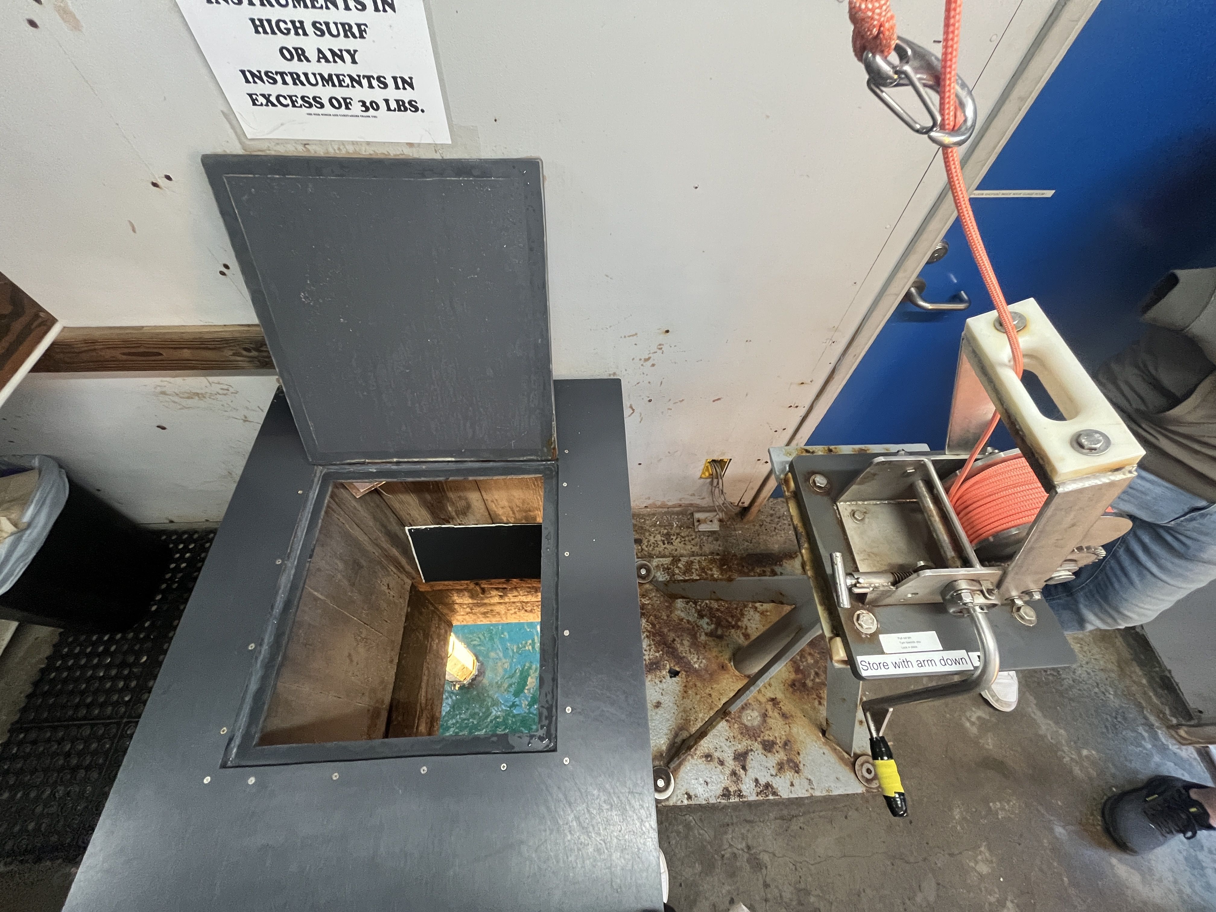 Open hatch revealing water below inside a room with worn walls, metal equipment with orange rope, and a sign partially visible about instrument weight limits.