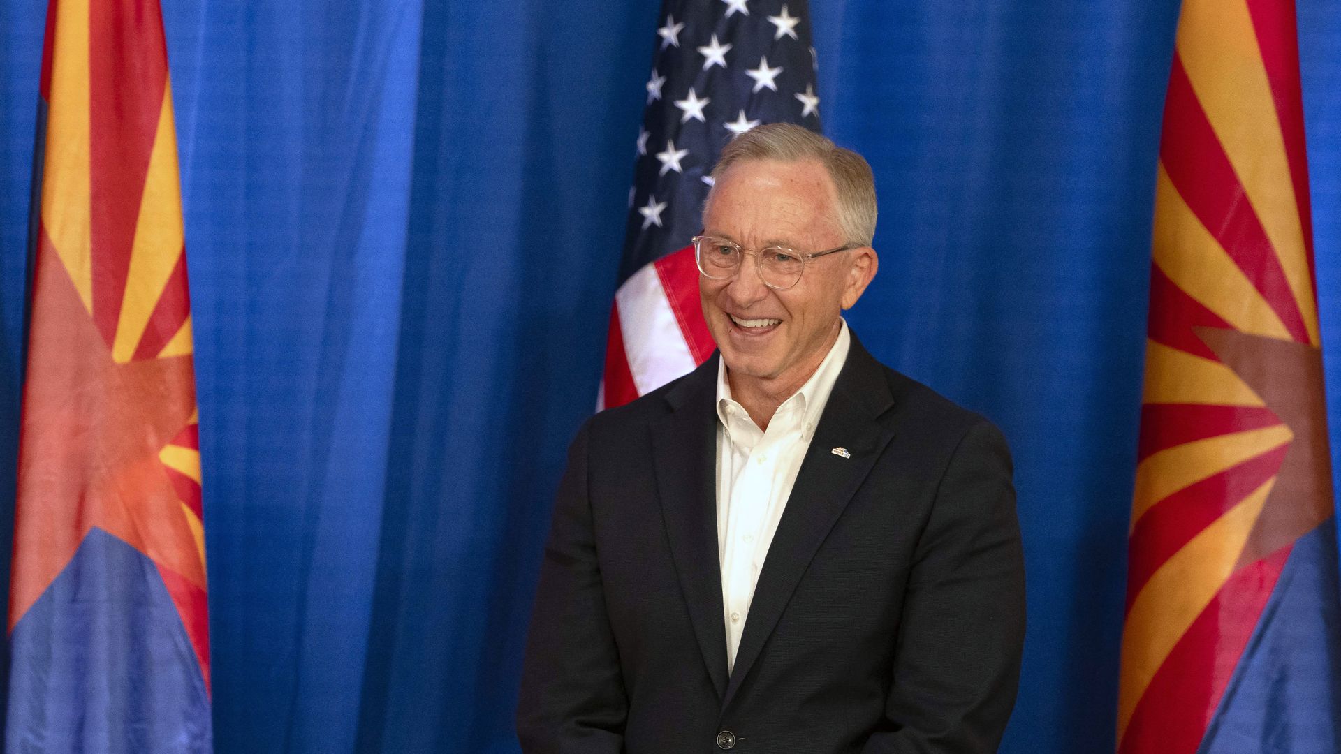 A smiling man in glasses wearing a black suit jacket over a white shirt stands in front of a blue background, with a United States flag directly behind him and Arizona flags to the left and right of it.