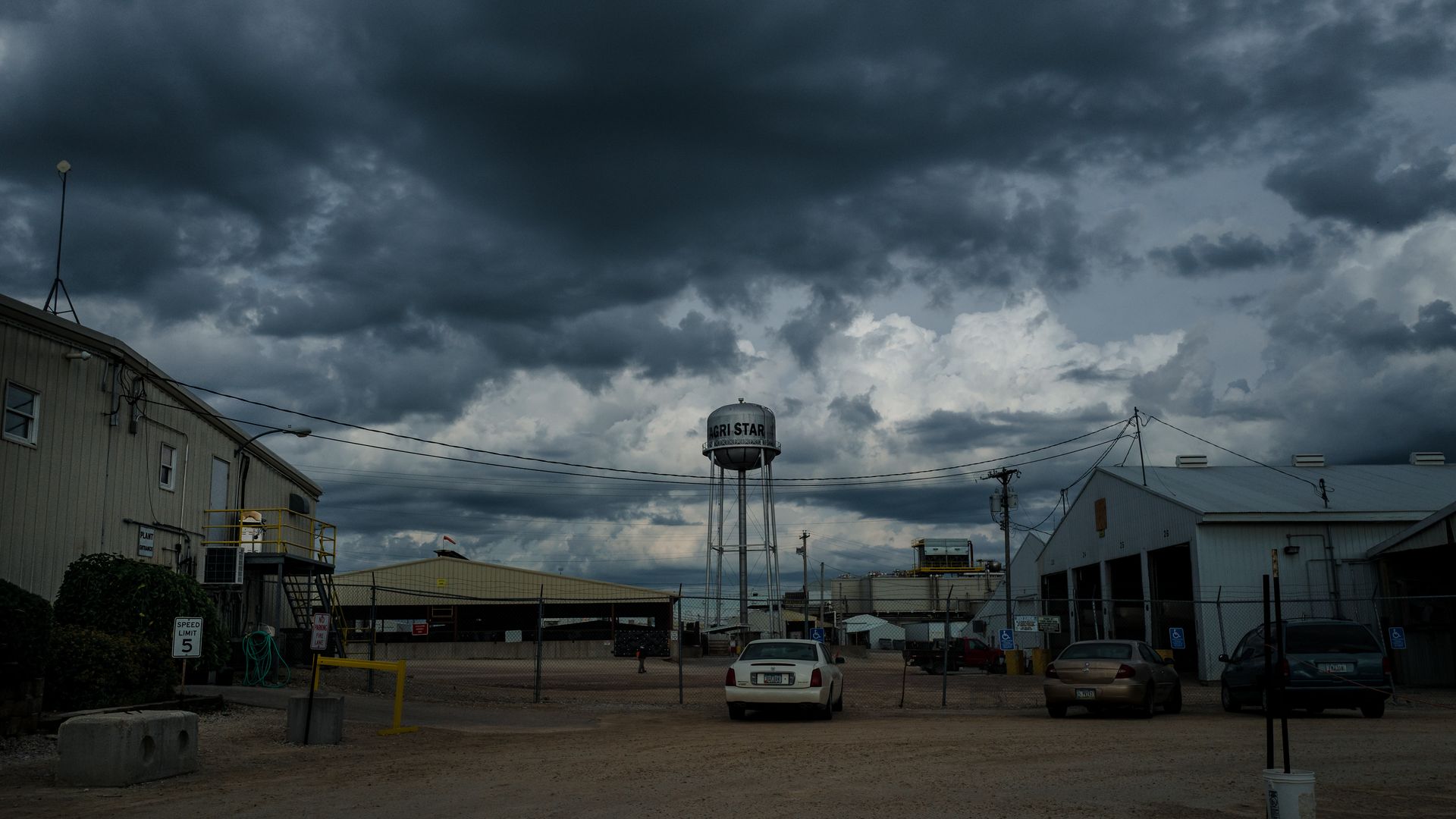 Dark clouds over an industrial site with a tall water tower labeled "AGRI STAR," several buildings, chain-link fences, and parked cars on a dirt ground.