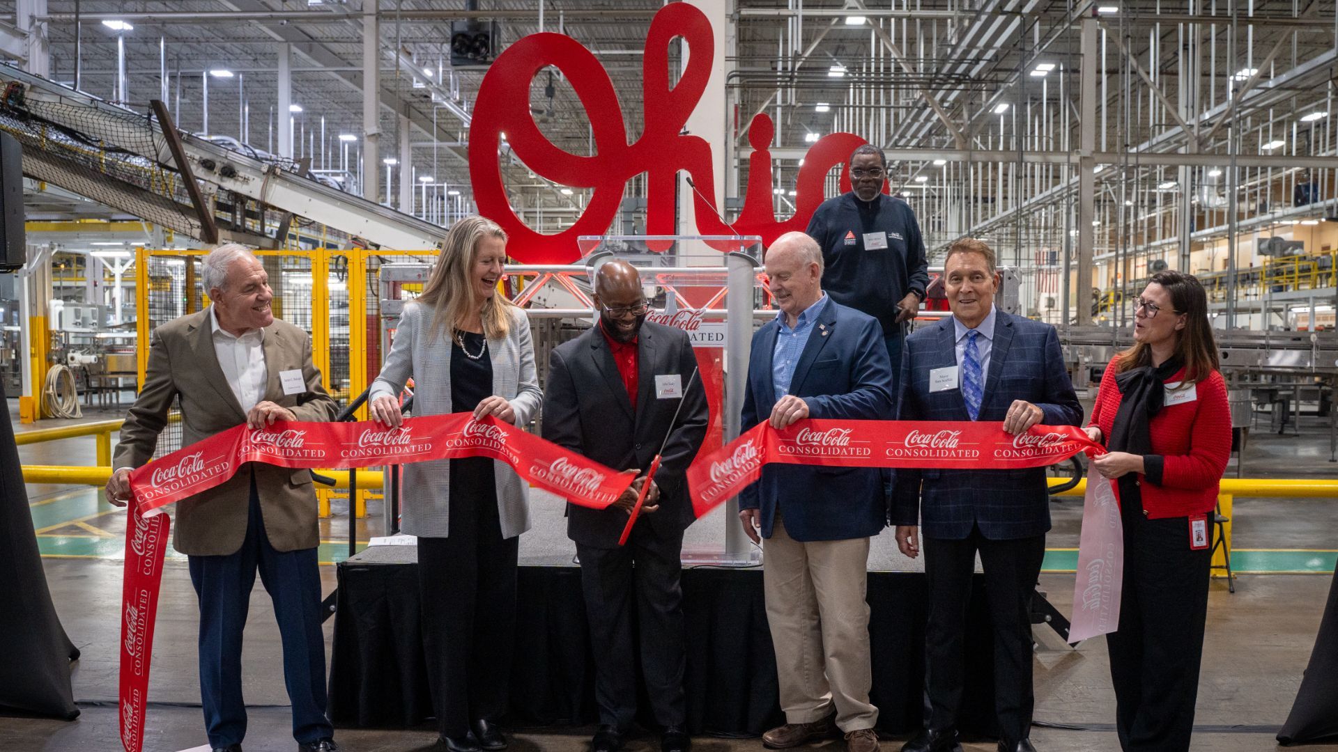 Group of six people, some holding a red Coca-Cola Consolidated ribbon, one cutting it, inside a large industrial facility with a big red "Ohio" sign in the background.