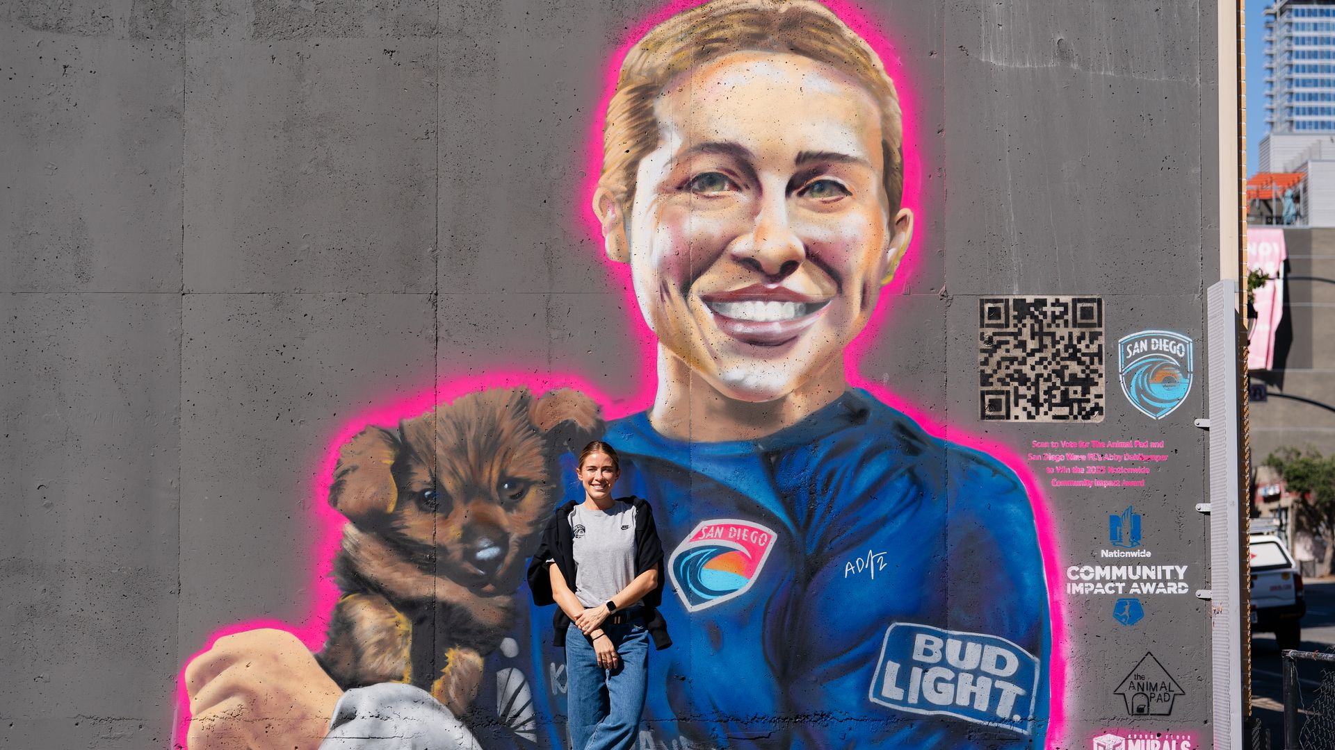 A woman stands in front of a mural depicting a San Diego Wave soccer player holding a puppy.