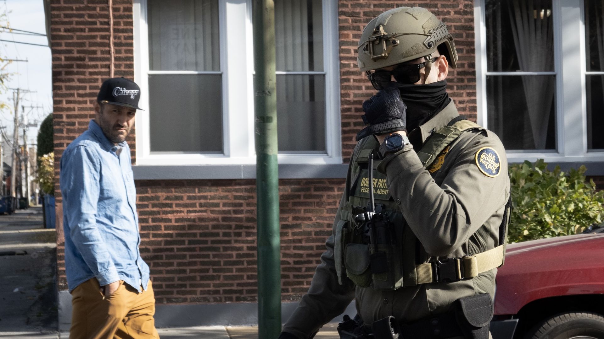 A civilian man in a light blue shirt and tan pants stands on a sidewalk beside a brick building as a heavily armored border patrol officer in a helmet and tactical gear stands nearby.