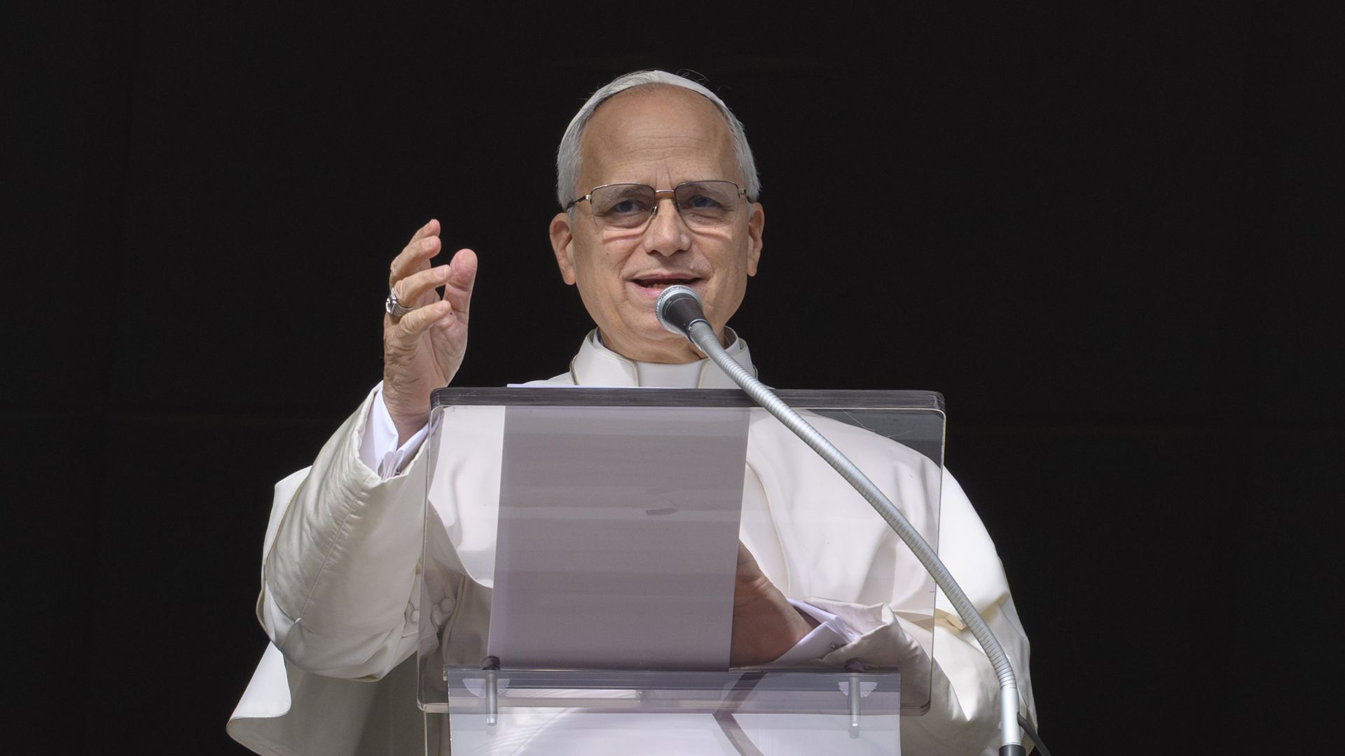 Pope Leo XIV, with glasses and short, gray hair and dressed in  white religious attire, speaks at a podium with a silver microphone against a black background.
