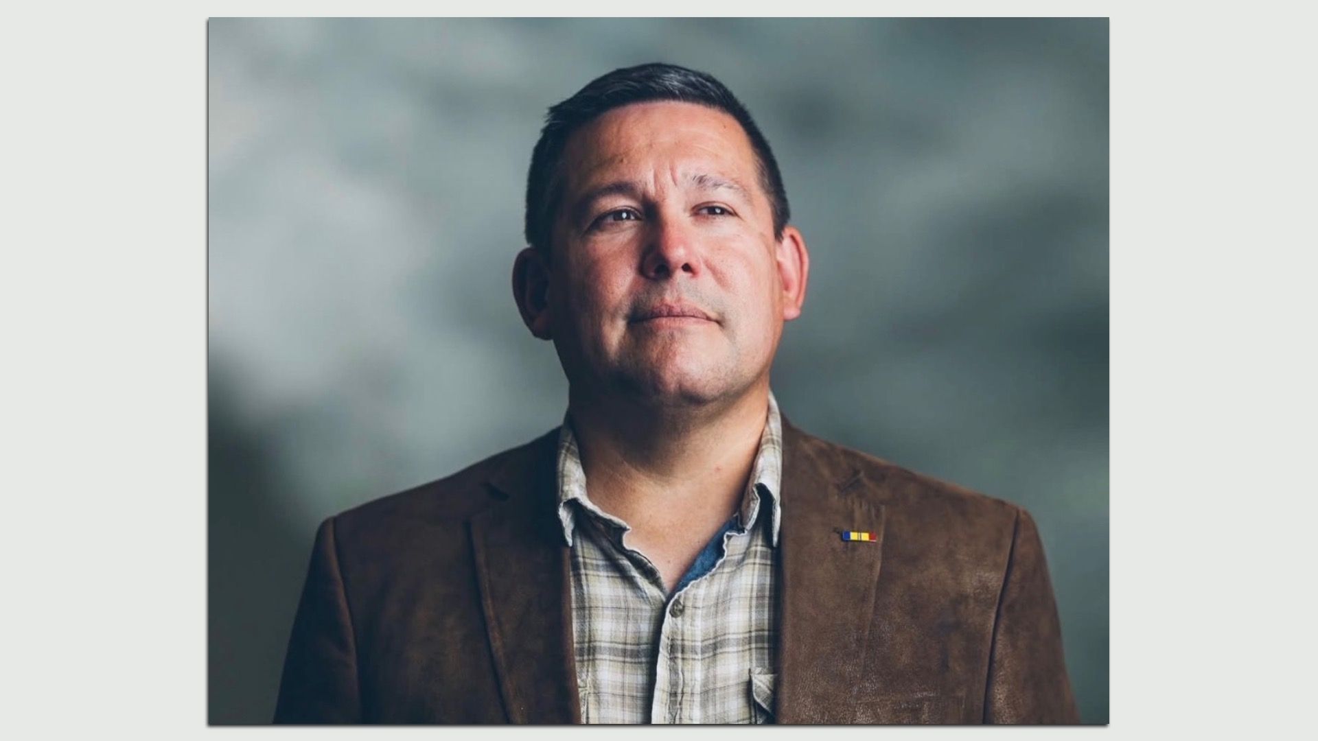 Portrait of a man with short dark hair, wearing a brown blazer over a plaid shirt, and a small multicolor lapel pin, against a soft gray-green blurred background.