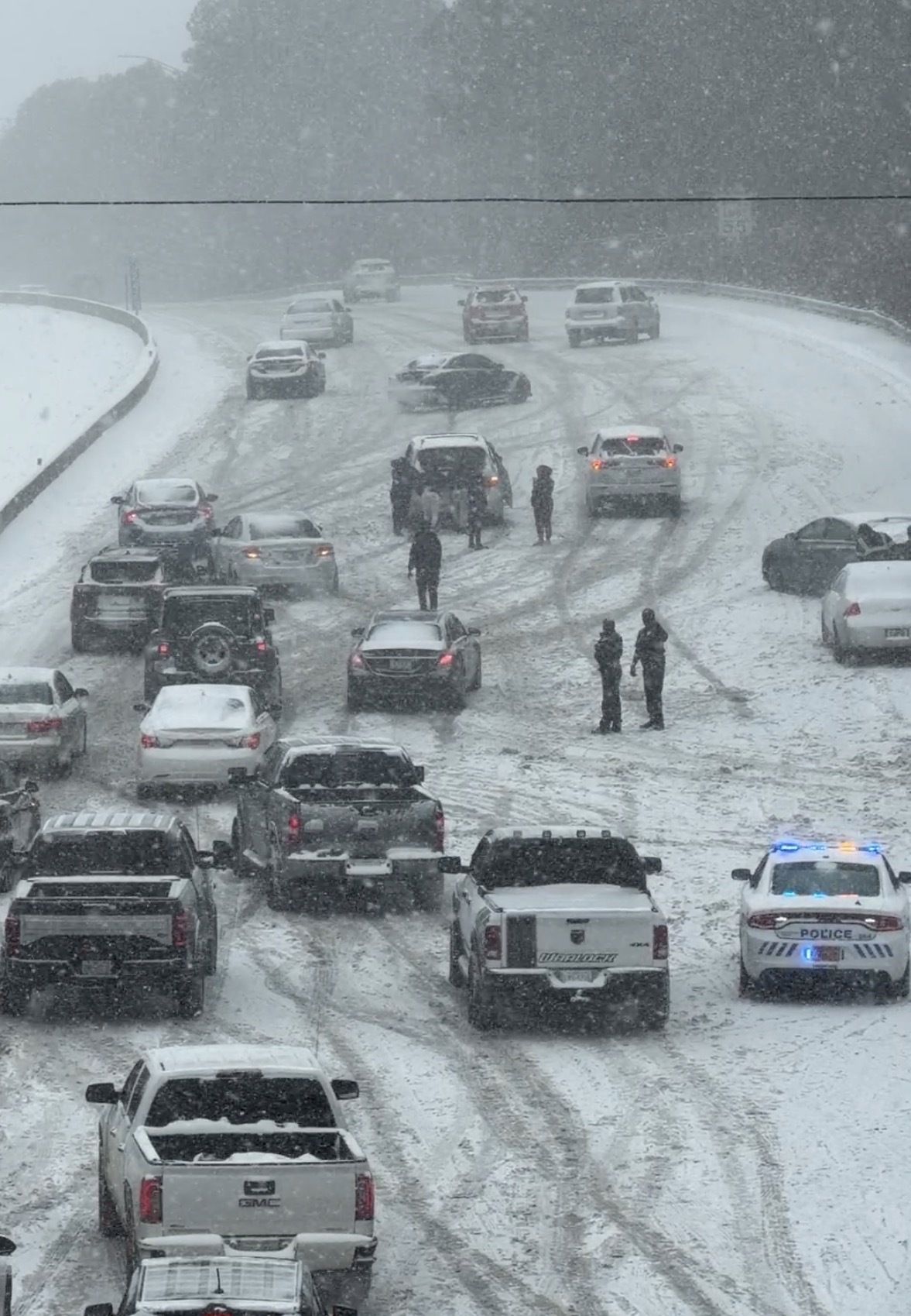 People pushing a car up a snow covered highway while several cars wait behind them. 
