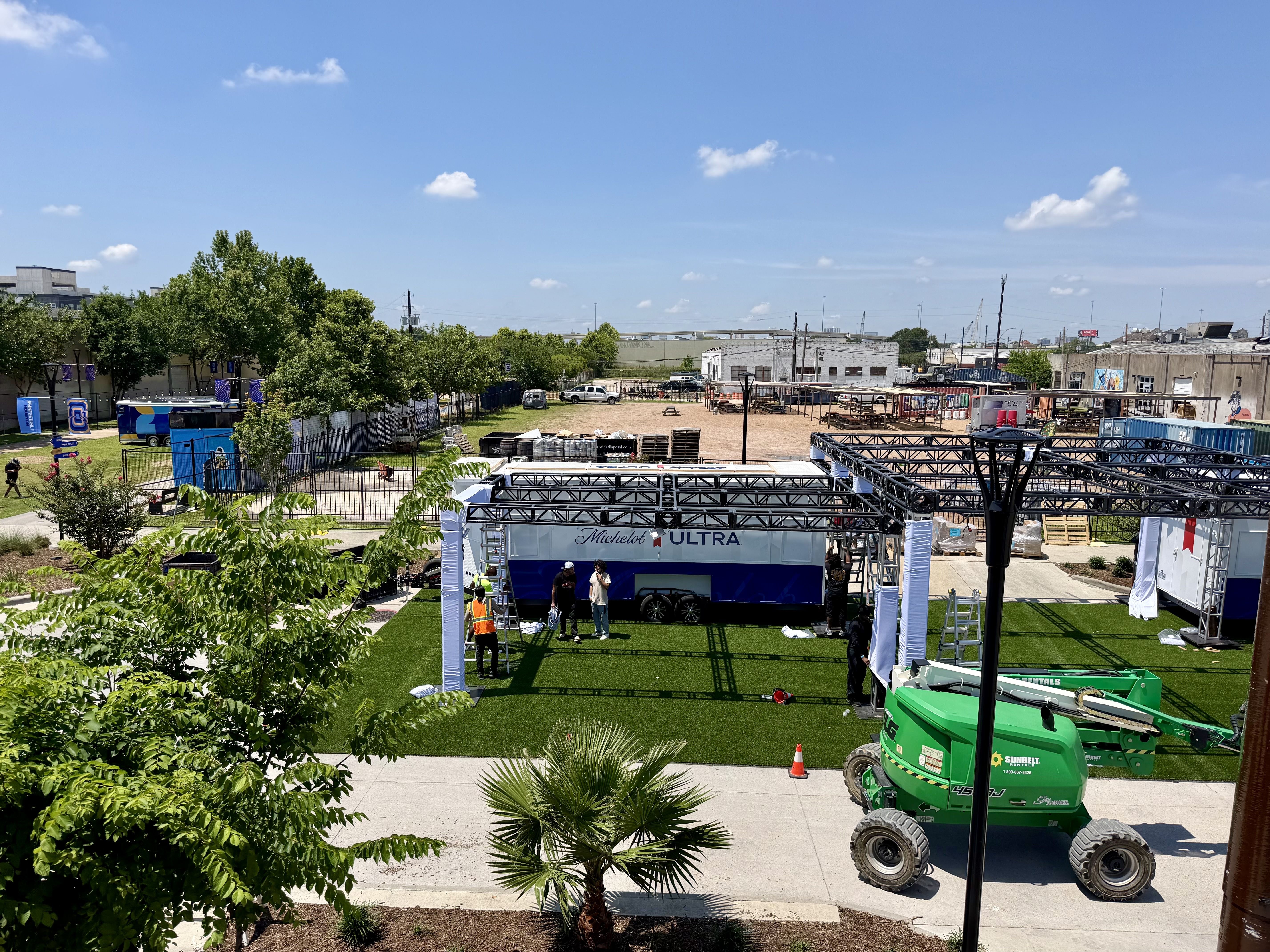 Outdoor event setup in a sunny, industrial yard. A blue and white Michelob Ultra trailer stage with metal scaffolding; workers in orange vests amid green trees and blue sky.