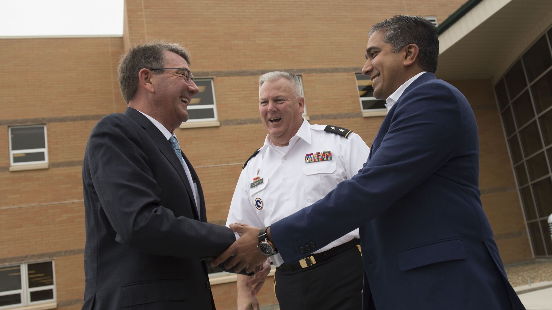 Ash Carter, in a black jacket, shakes hands with Raj Shah, in a blue jacket. A man in the middle is wearing white. A brick building is seen behind them.