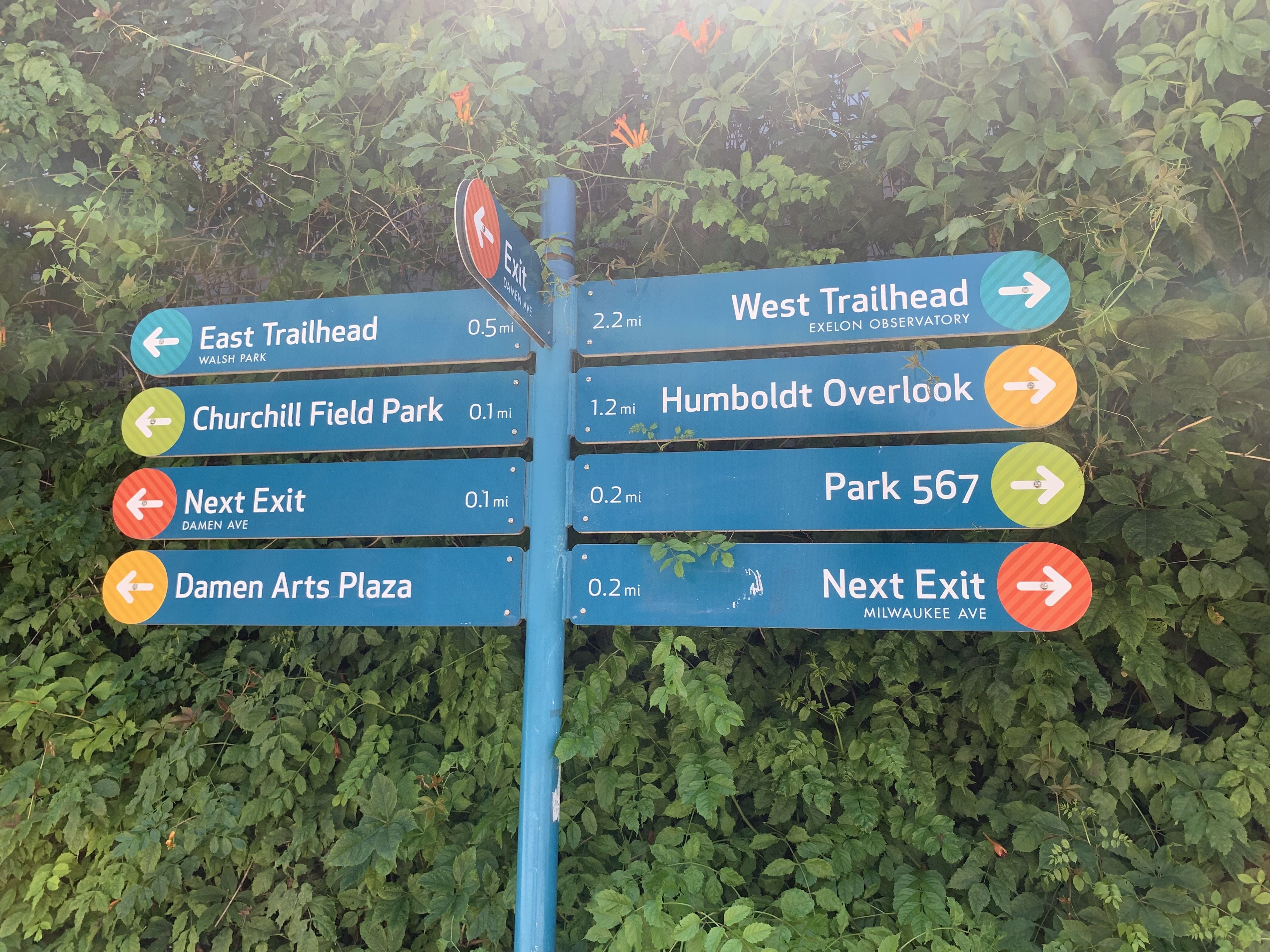 Blue signpost with multiple trail signs—East Trailhead, West Trailhead, Churchill Field Park, and Humboldt Overlook—set among dense green ivy; a leaning orange "Exit" sign nearby.