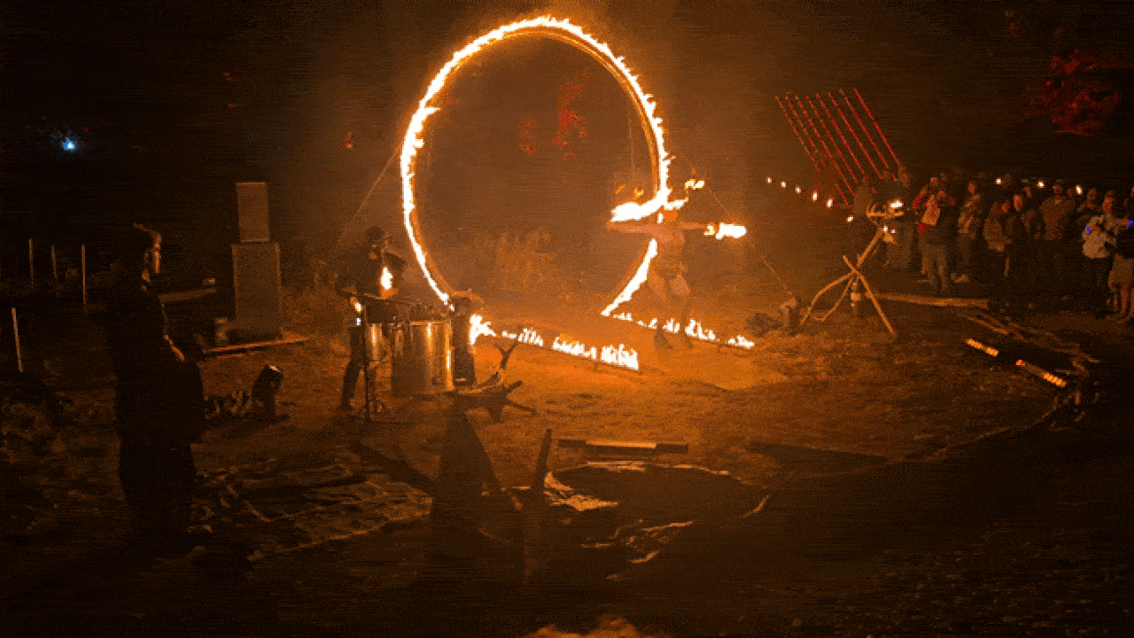 Nighttime fire performance with a performer spinning a flaming hoop and others playing steel drums, surrounded by a crowd watching the fiery spectacle outdoors.