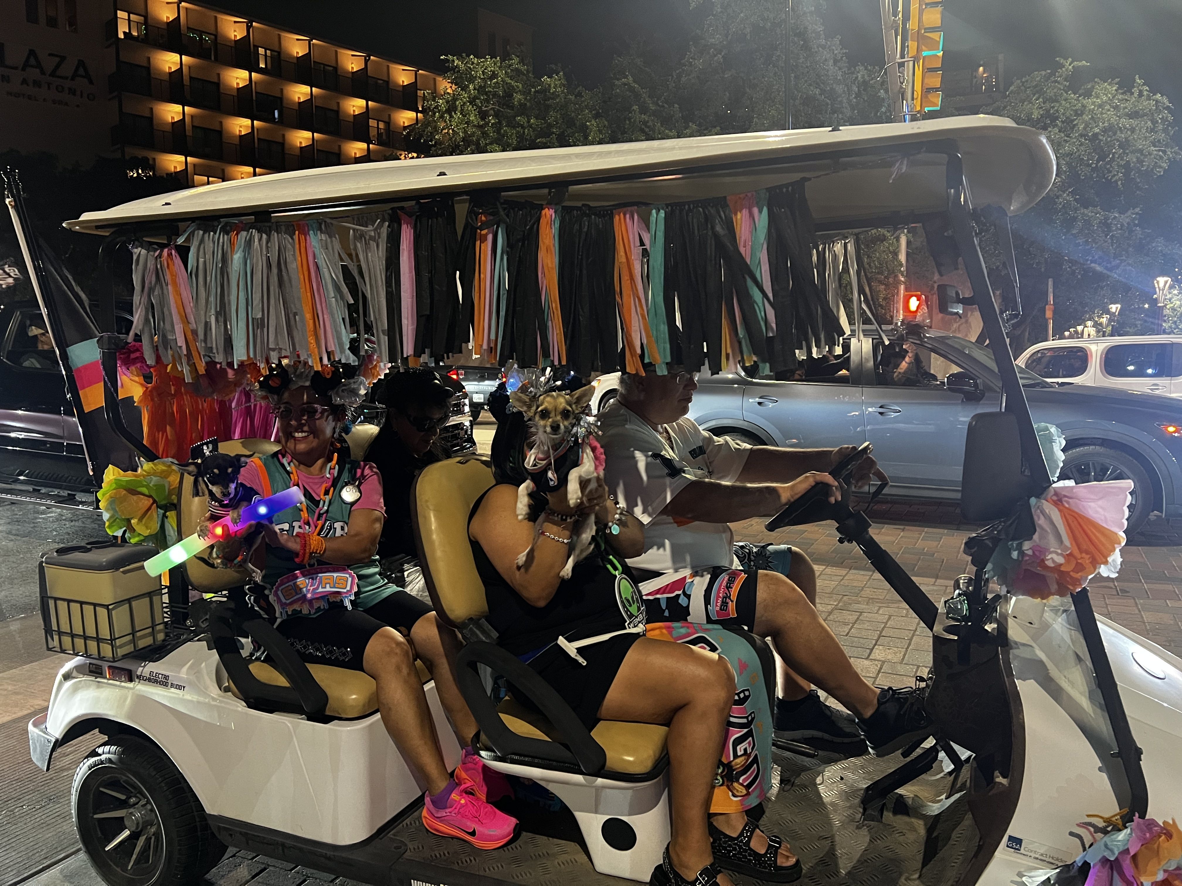Nighttime party bus with colorful tassels along the open roof; two riders in bright outfits, two small dogs on laps, glow sticks, and a city street with lit buildings in the background.