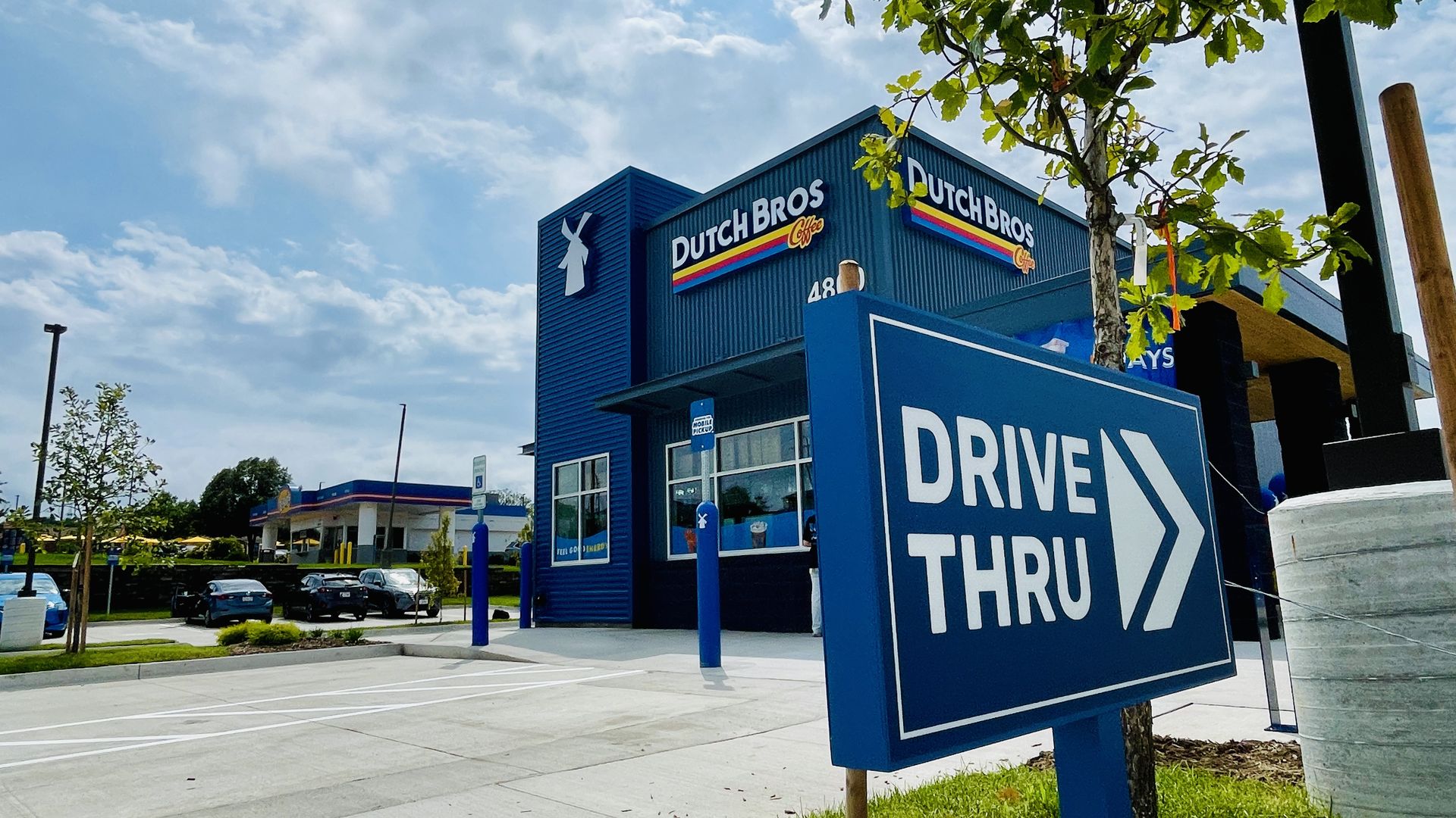Blue Dutch Bros Coffee building with a white windmill logo and a striped sign; a large blue DRIVE THRU sign dominates the foreground as a tree and clear sky frame the scene.