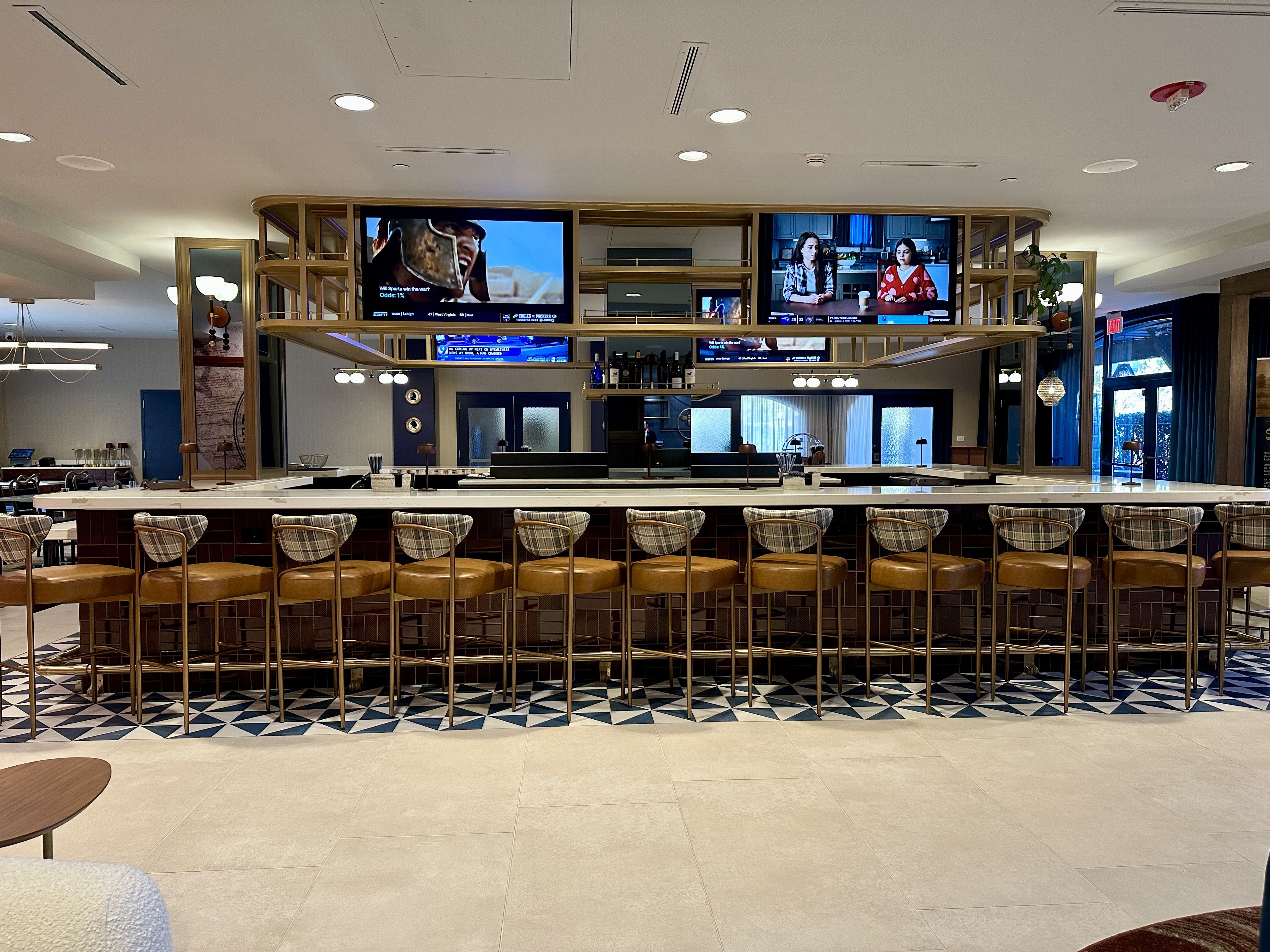 Modern bar with a long marble countertop and 12 tan leather stools with plaid backs, tiled floor area, two TVs above the bar displaying a warrior and two women, ambient lighting.
