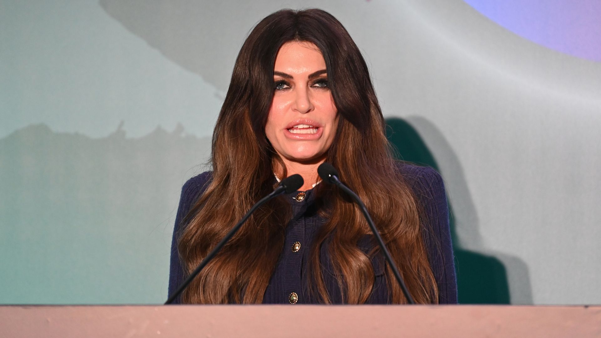 Kimberly Guilfoyle, with long dark hair speaks at a podium with several microphones on a stage with a pink edge, wearing a dark blazer. A pastel backdrop with purple shapes behind her.