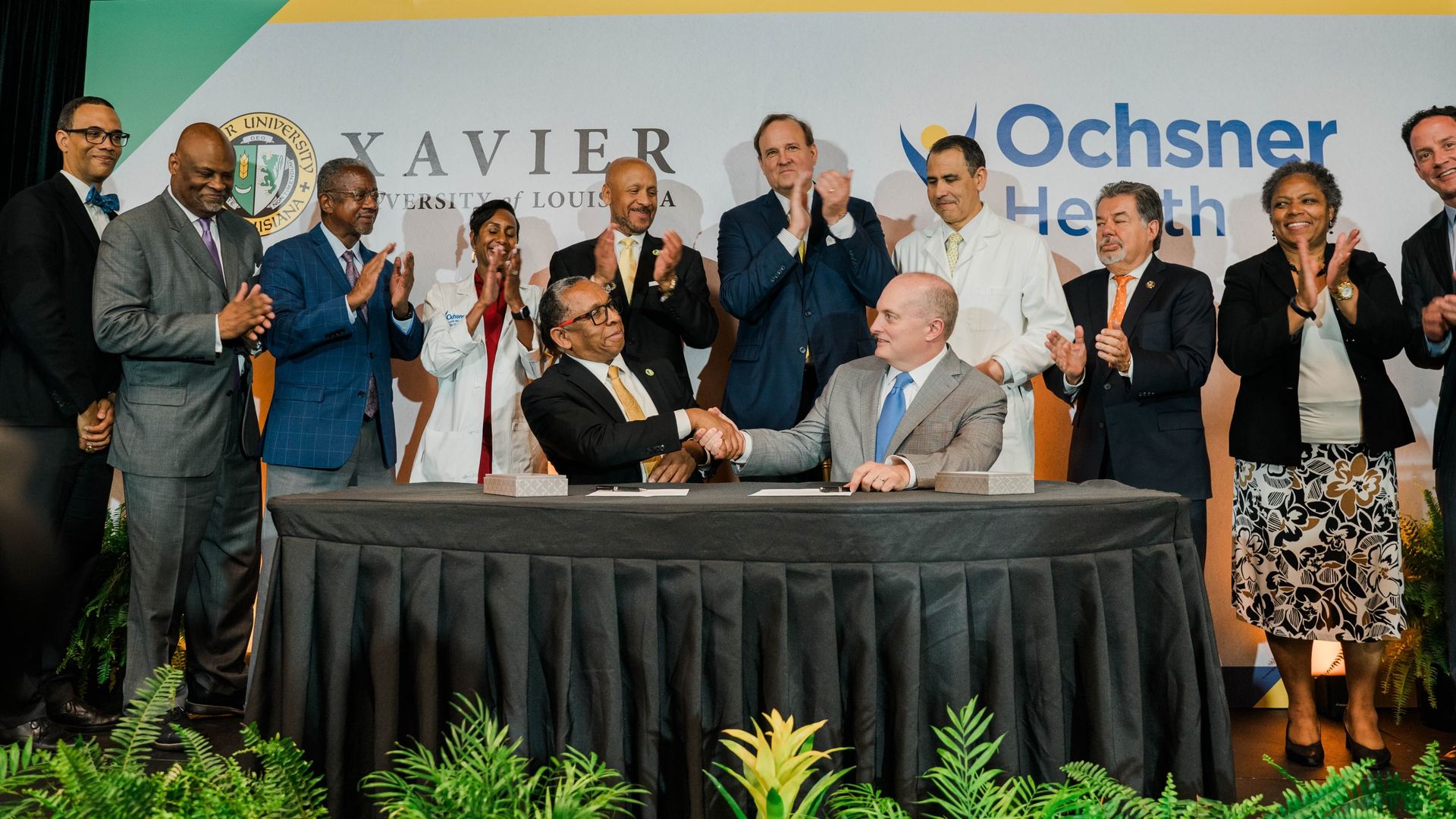 Photo shows two men shaking hands at a table with a sign behind them for Xavier University and Ochsner Health.