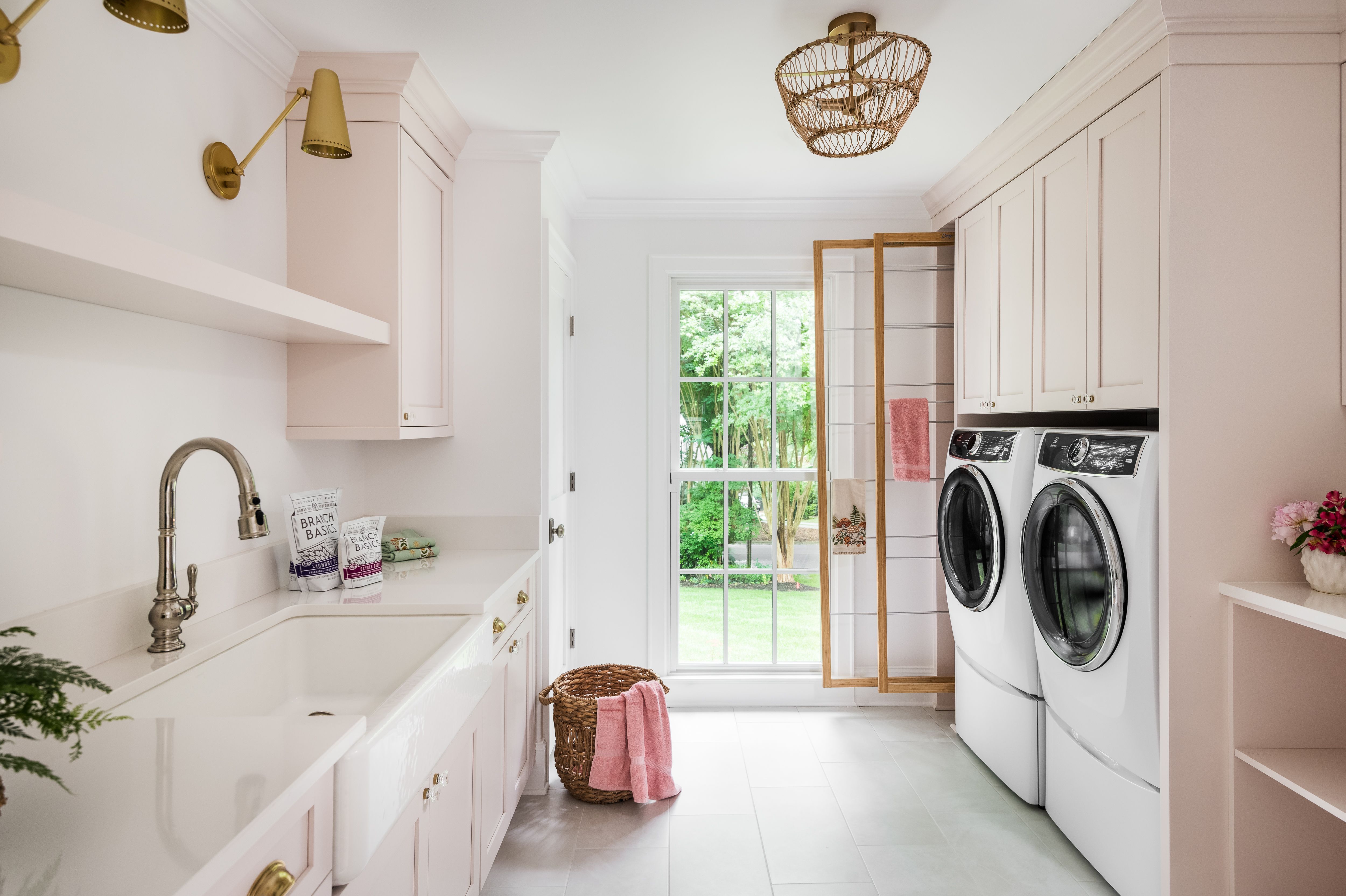 Bright laundry room with white and pastel pink cabinets, white front-load washer and dryer, farmhouse sink, woven basket with pink towel, and large window showing green outdoor trees.