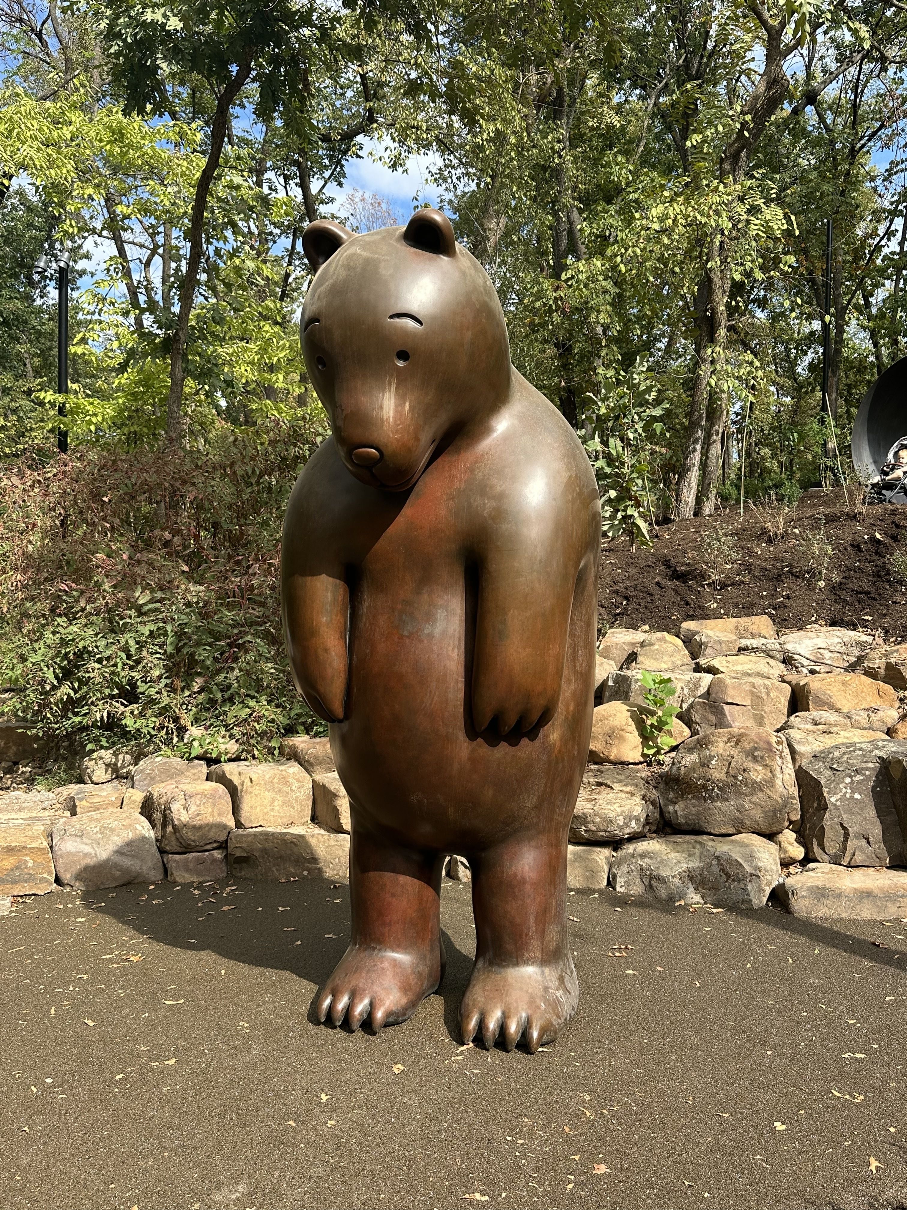 Bronze bear statue in a park, standing upright with rounded body and oversized paws. Surrounded by trees, a rocky wall, and a clear blue sky.