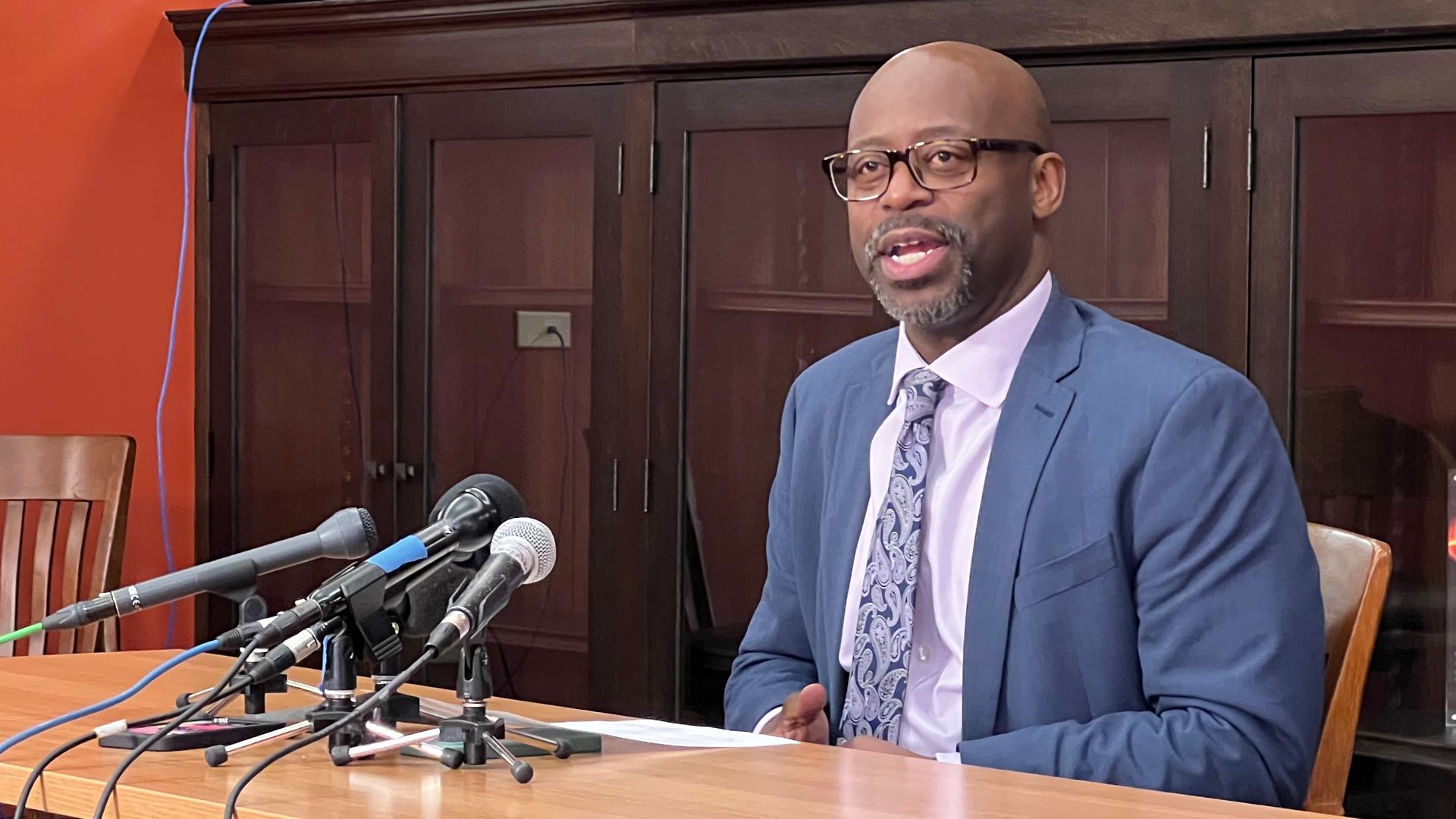 A man in a blue suit and patterned tie sits at a wooden table with multiple microphones in front of him, addressing an unseen audience. An orange-walled room with dark cabinets behind him.
