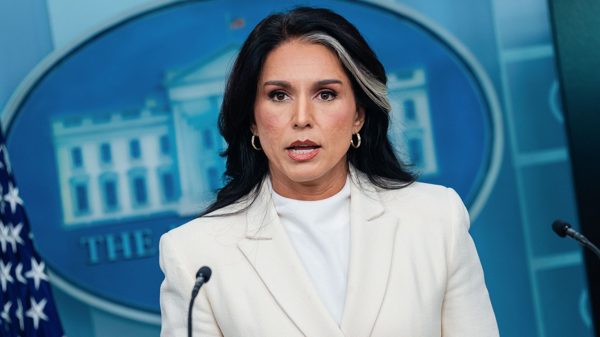 Tulsi Gabbard, director of National Intelligence, wearing black hair and white streak, and a white blazer, speaking at podium with microphones in front of a blue background featuring the White House emblem and an American flag.