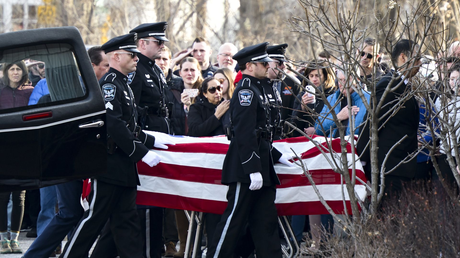 An honor guard of uniformed police officers carries a casket draped in an American flag out of a hearse as a crowd looks on.