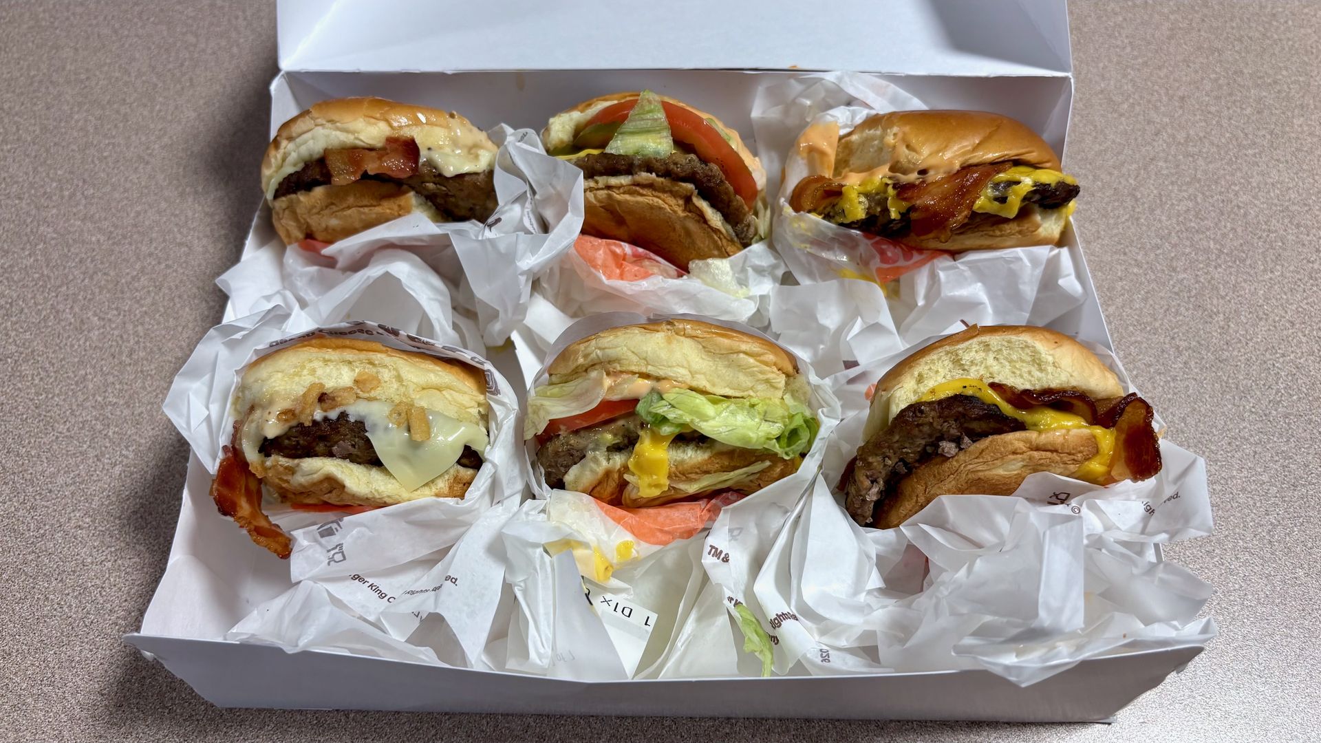 Open white takeout box with six burgers wrapped in paper on a speckled beige counter. Buns, beef patties, melted cheese, lettuce, tomato, bacon, and various sauces are visible on several burgers.