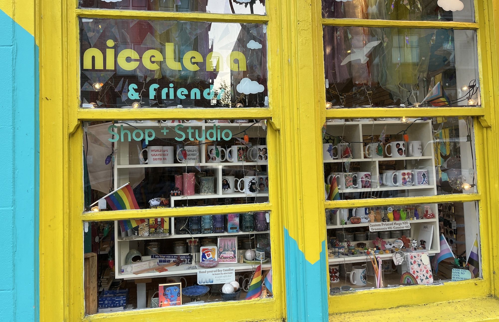 Bright yellow and blue shopfront window of nicelena & friends Shop + Studio displaying mugs, candles, pride flags, ornaments, and colorful small items inside.