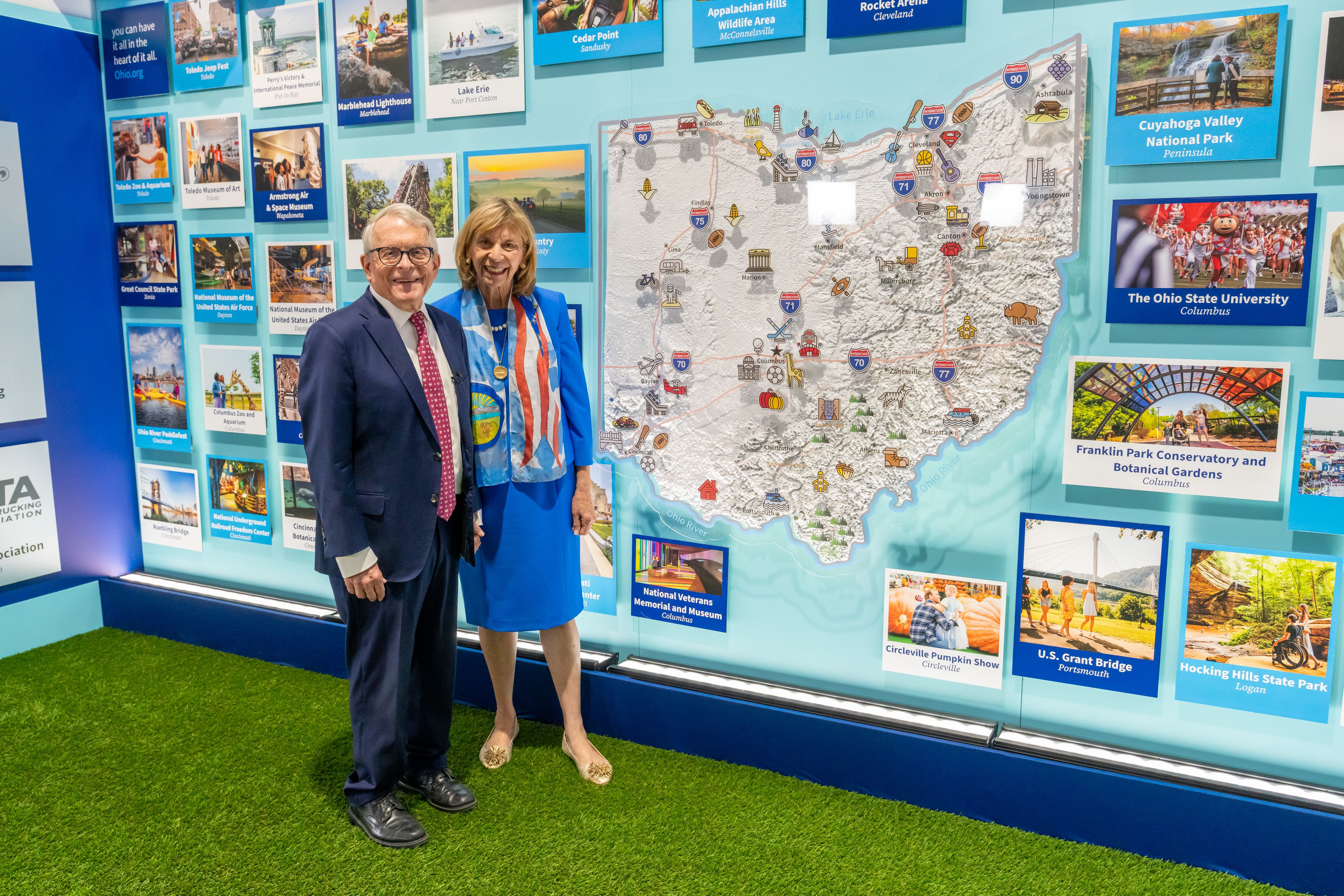 Gov. Mike DeWine and First Lady Fran DeWine pose in front of a Great American State Fair exhibit filled with photo cards and a large, illustrated map of Ohio showing parks and landmarks.