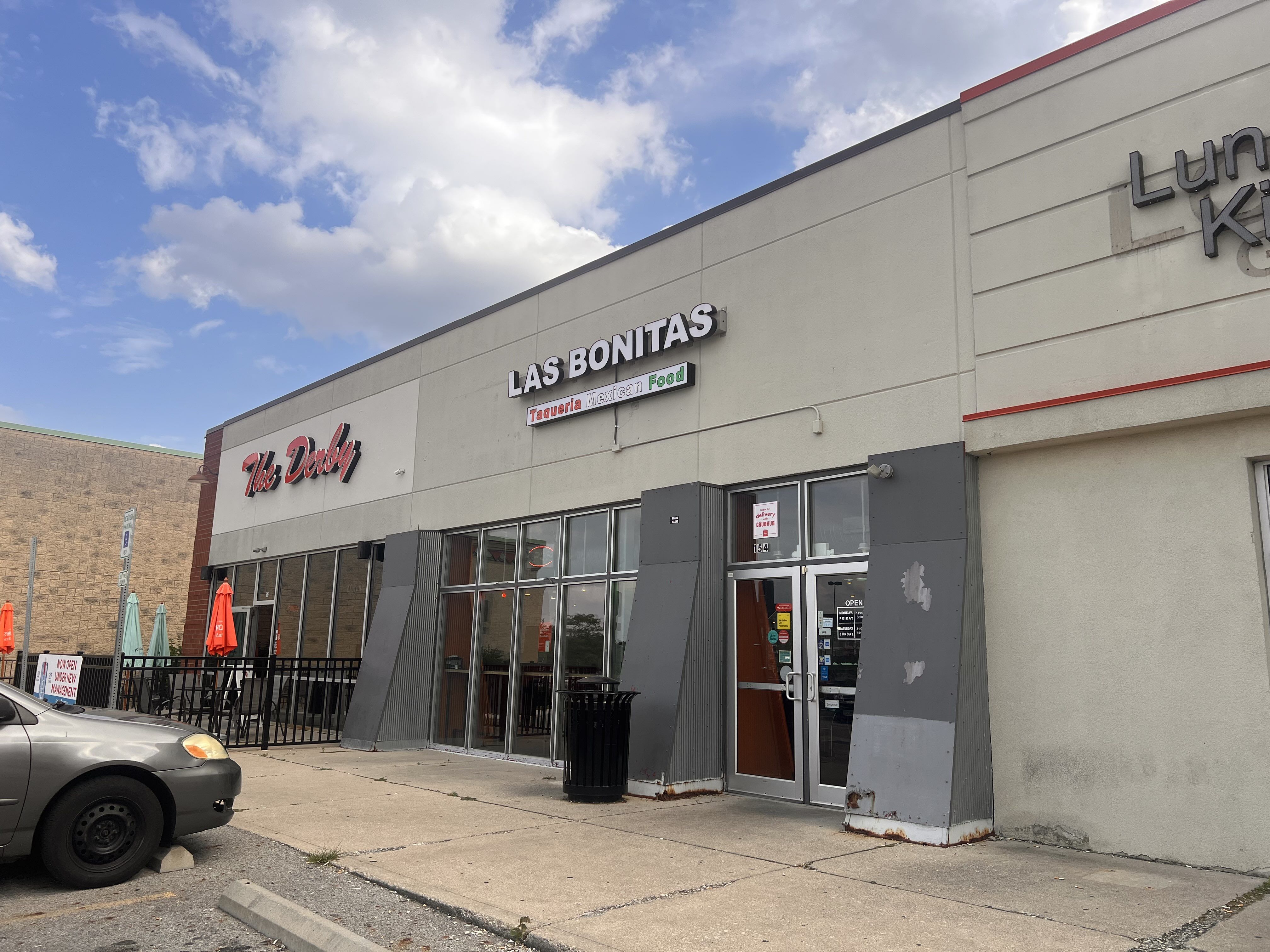 Exterior of a commercial building with two restaurants, The Derby with red signage and Las Bonitas Taqueria Mexican Food with white signage. Cars, patio tables, umbrellas, and partly cloudy sky.