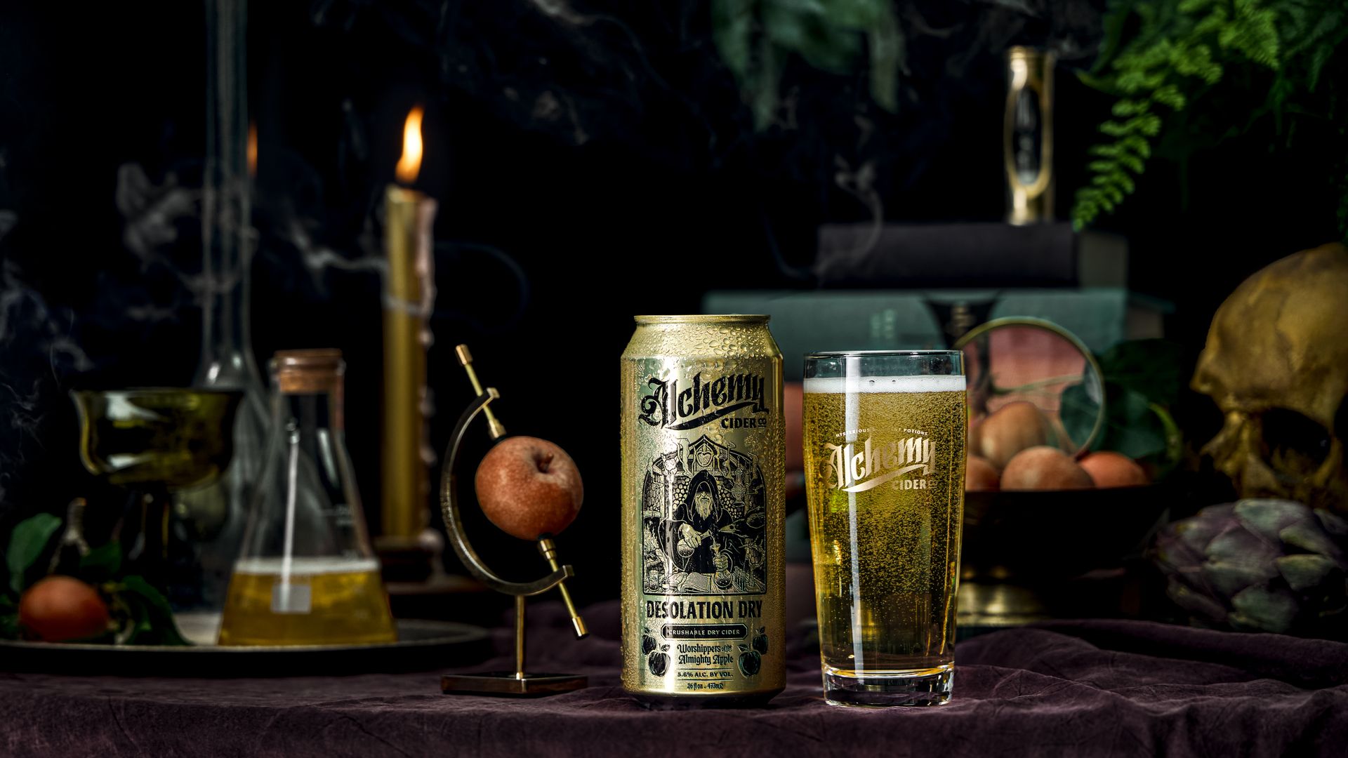 Gold can and glass of Alchemy Cider Desolation Dry with apple ornament, lit candle, flask, skull, and dark leafy background on purple cloth.