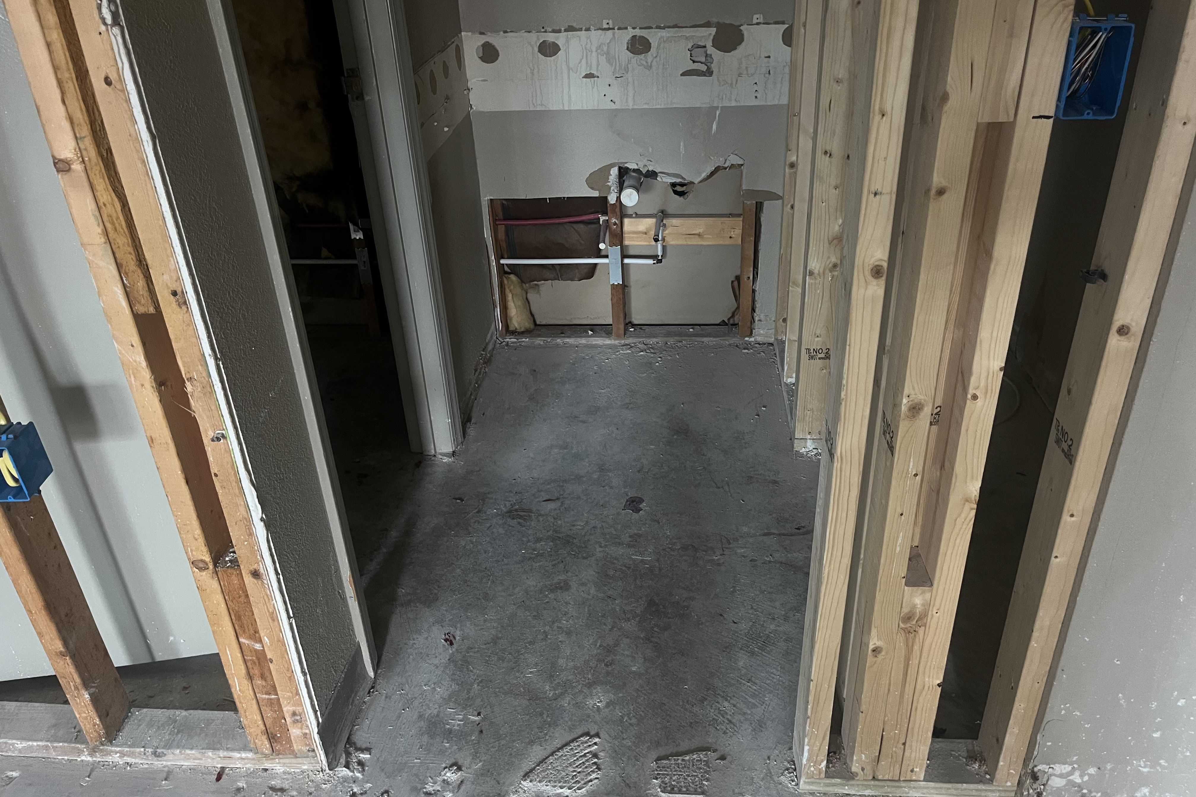 Room under renovation with exposed wooden studs, concrete floor, and partially removed drywall revealing plumbing and insulation.
