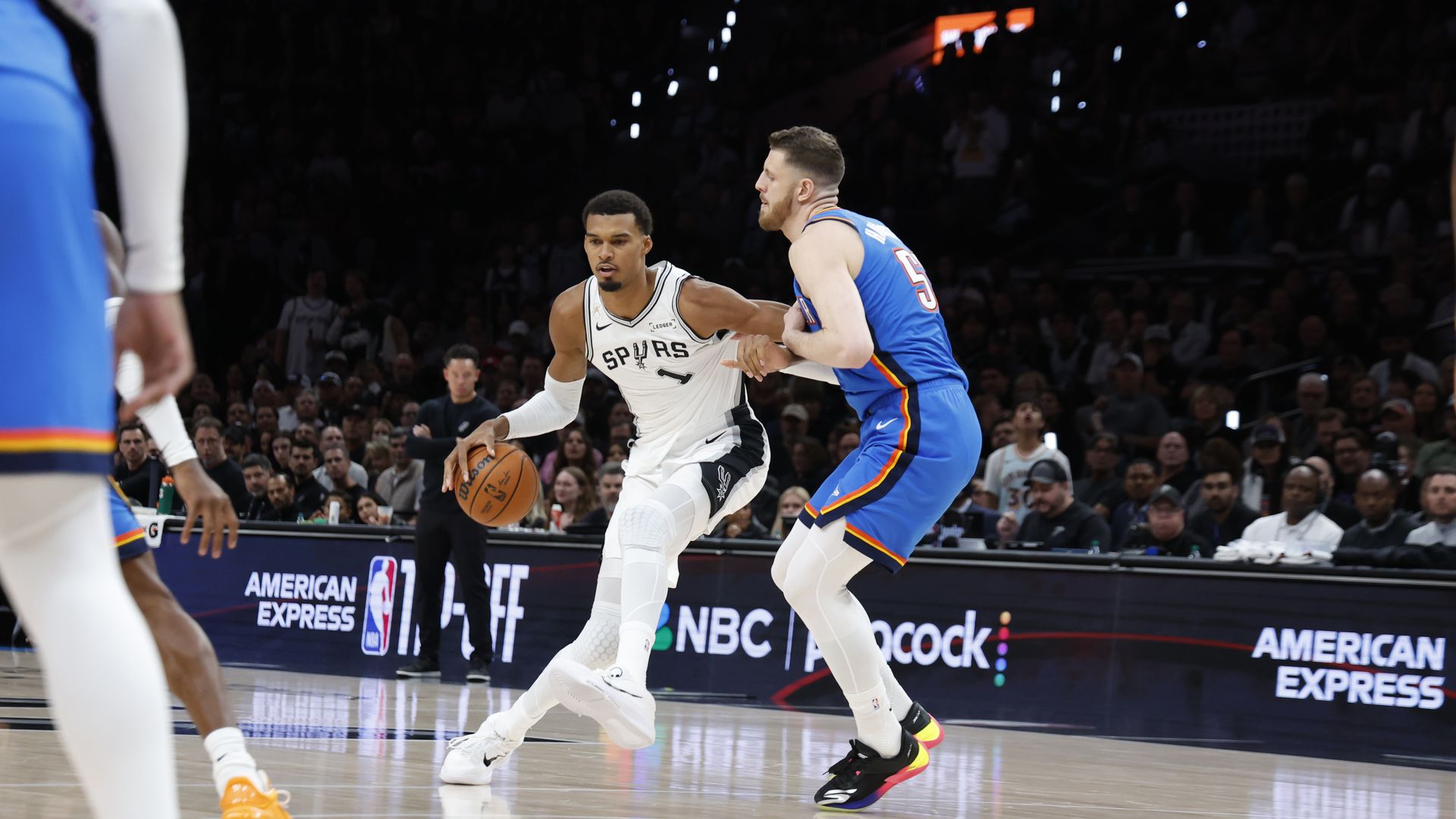 Spurs player in white dribbles the ball as a Thunder defender in blue guards him; crowded arena with illuminated ads along the sideline.