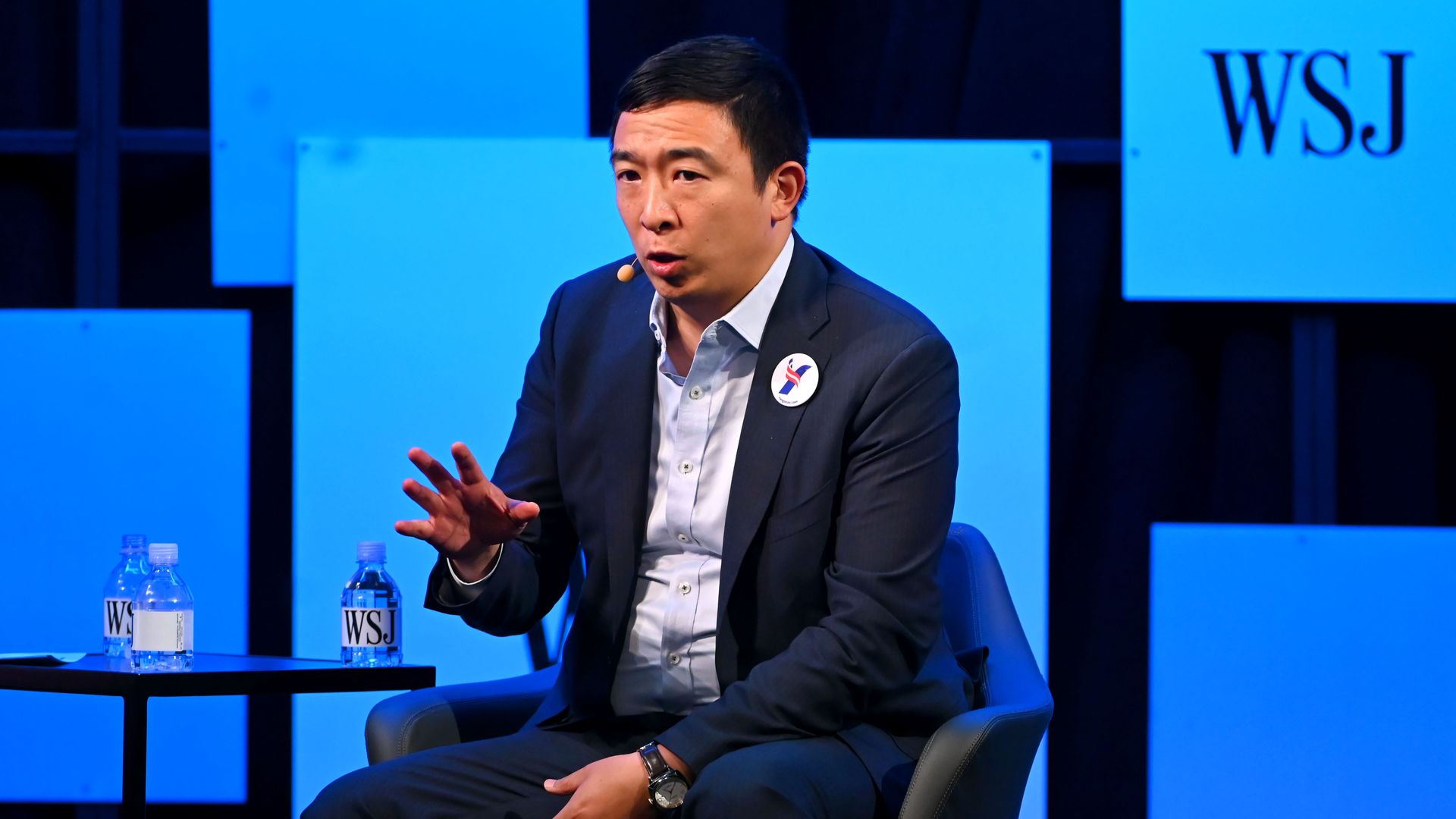 Andrew Yang sitting in a chair while extending his hand forward. he is wearing a suit, Yang button and in front of some Wall Street Journal graphics. 
