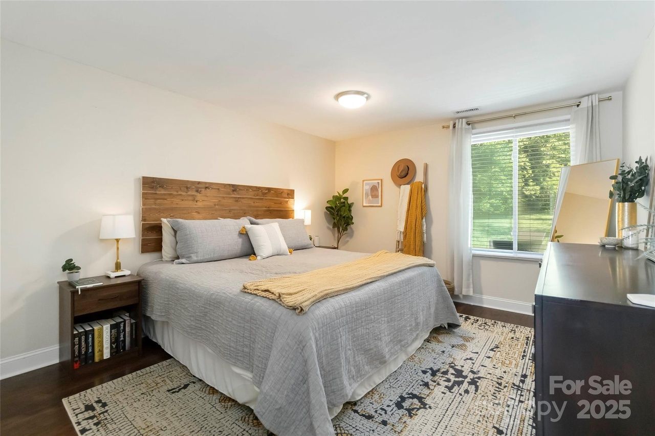 Bedroom with a large bed featuring a wooden headboard, gray bedding, and a yellow throw. Nightstand with lamp and books, window with white curtains, rug, and black dresser with mirror.