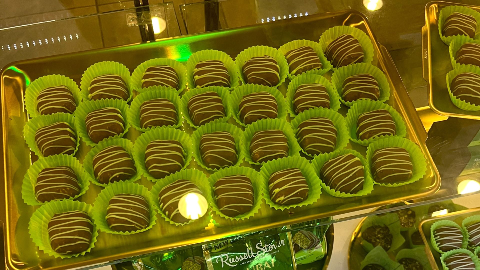 Tray of round chocolate truffles with white drizzle, each in green paper cups, displayed in a gold-colored tray under warm lighting.