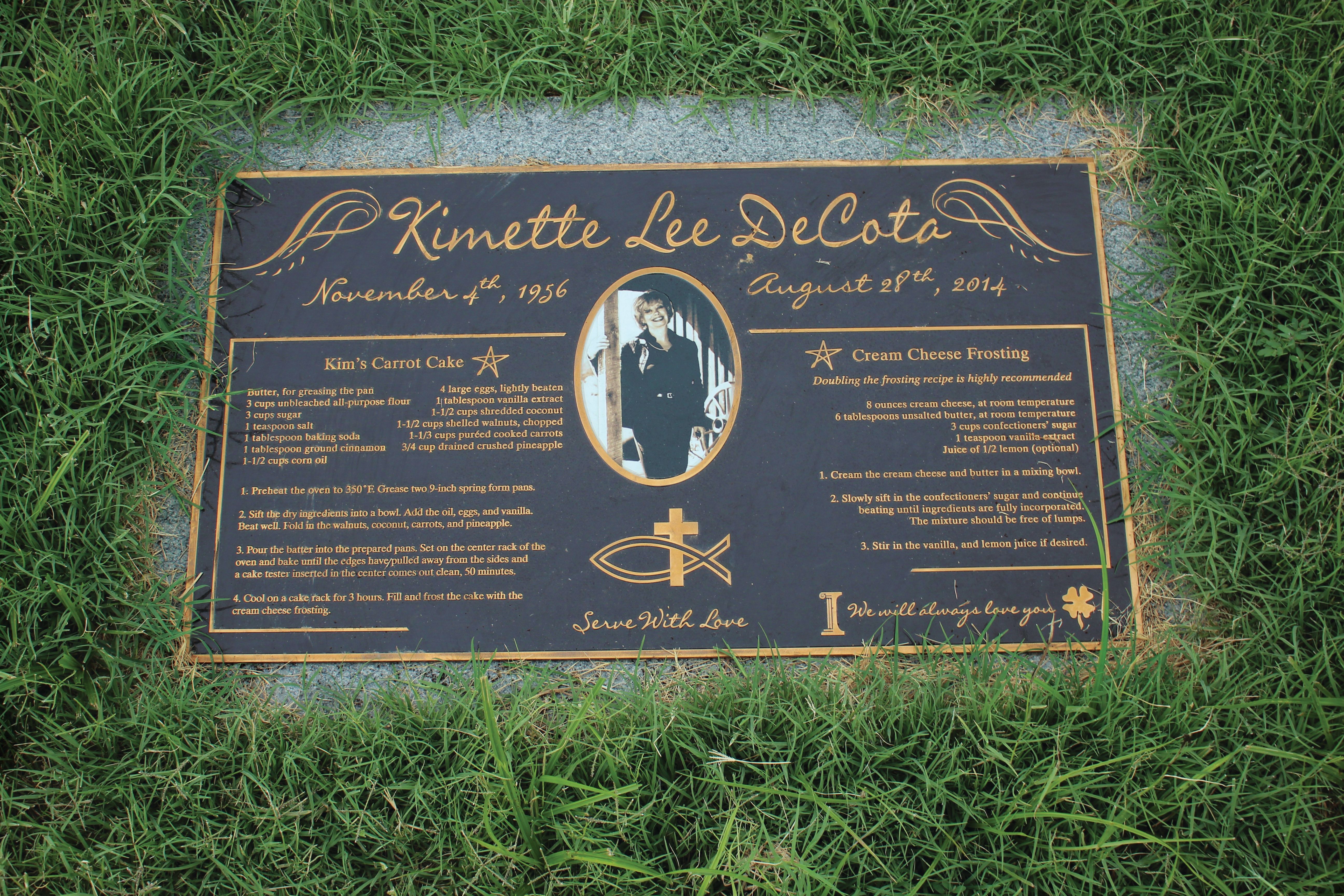 Memorial plaque for Kimette Lee DeCota (1956-2014) with her photo and a carrot cake recipe including cream cheese frosting, surrounded by grass.