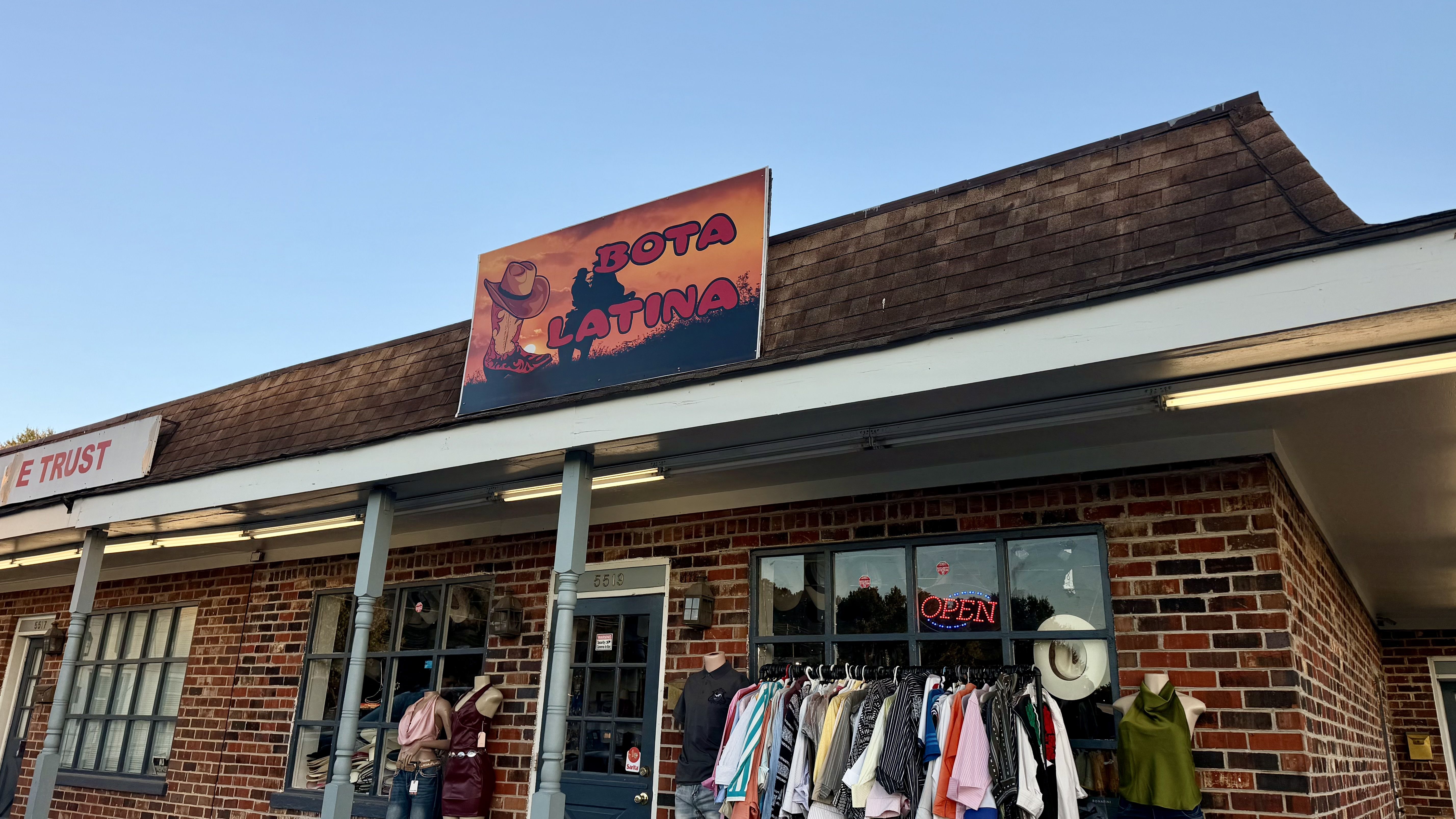 Brick storefront with brown shingle roof. A sign reads "BOTA LATINA" beside a cowboy boot image. Clothes racks and mannequins sit outside; a neon OPEN sign glows in the window.
