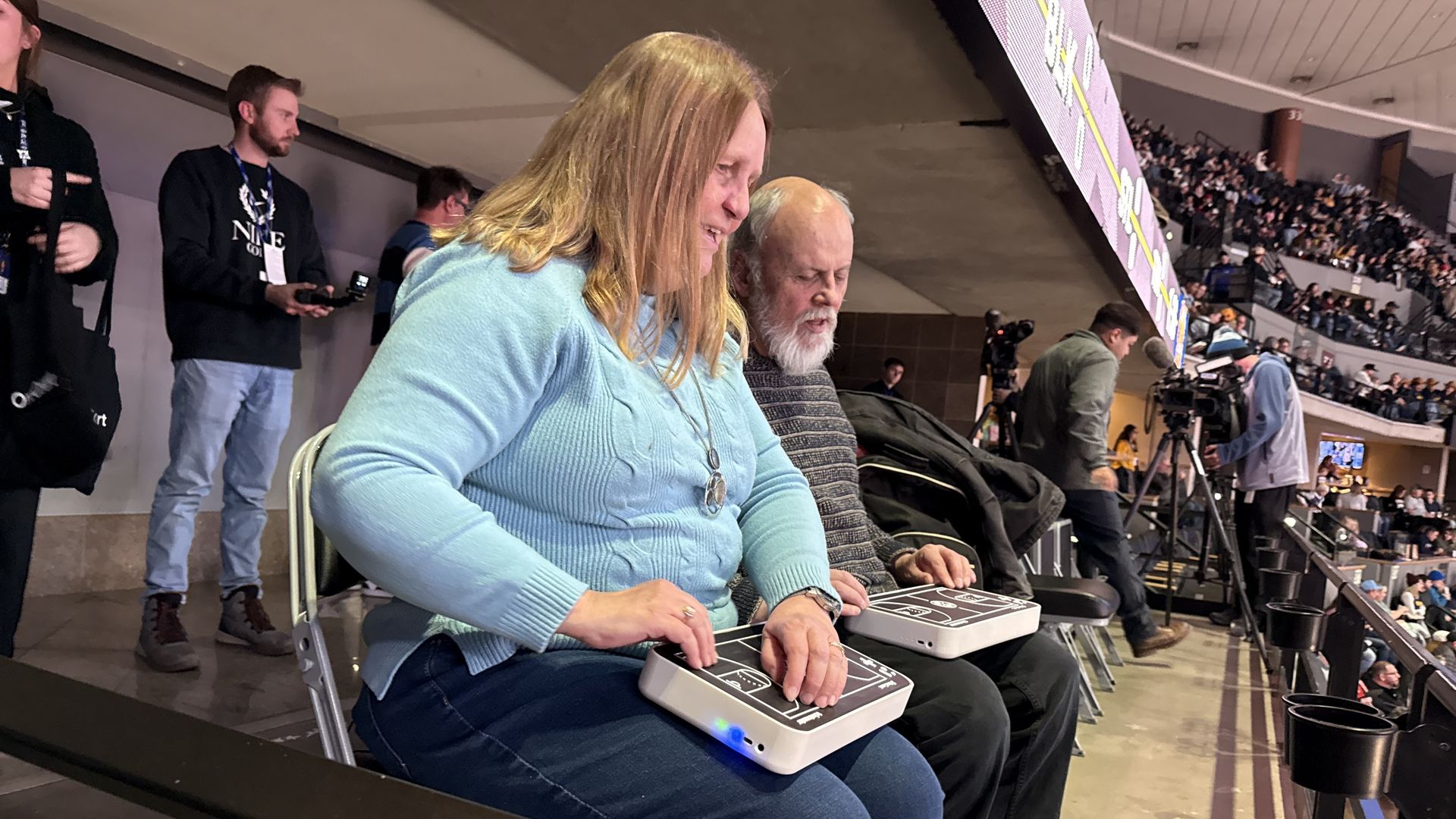 Two people sitting in a crowded arena, each using a device with a basketball court diagram, likely for audio description at a sports event, surrounded by other attendees and cameras.