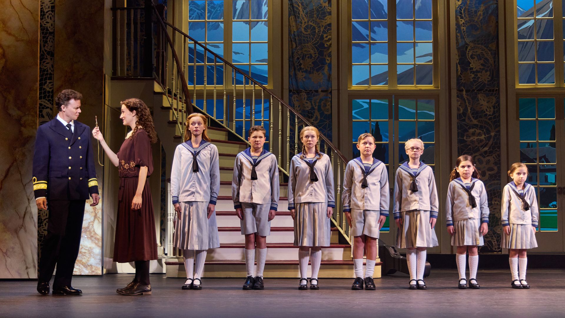 A man in a navy uniform and a woman in a brown dress stand beside seven children in blue-and-white sailor outfits on stage with a backdrop of tall windows and mountains.