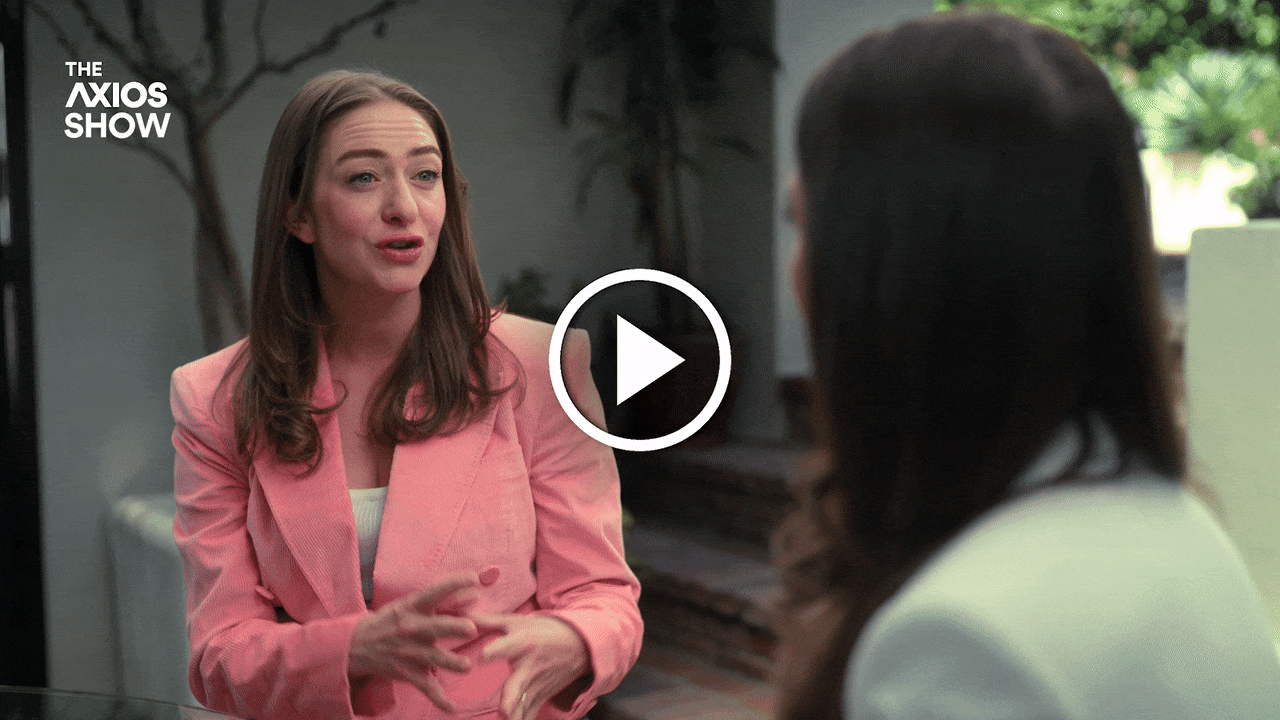 Outdoor interview: a woman in a pink blazer speaks and gestures, facing another woman with long dark hair. The Axios Show logo is top-left with a play button overlay.