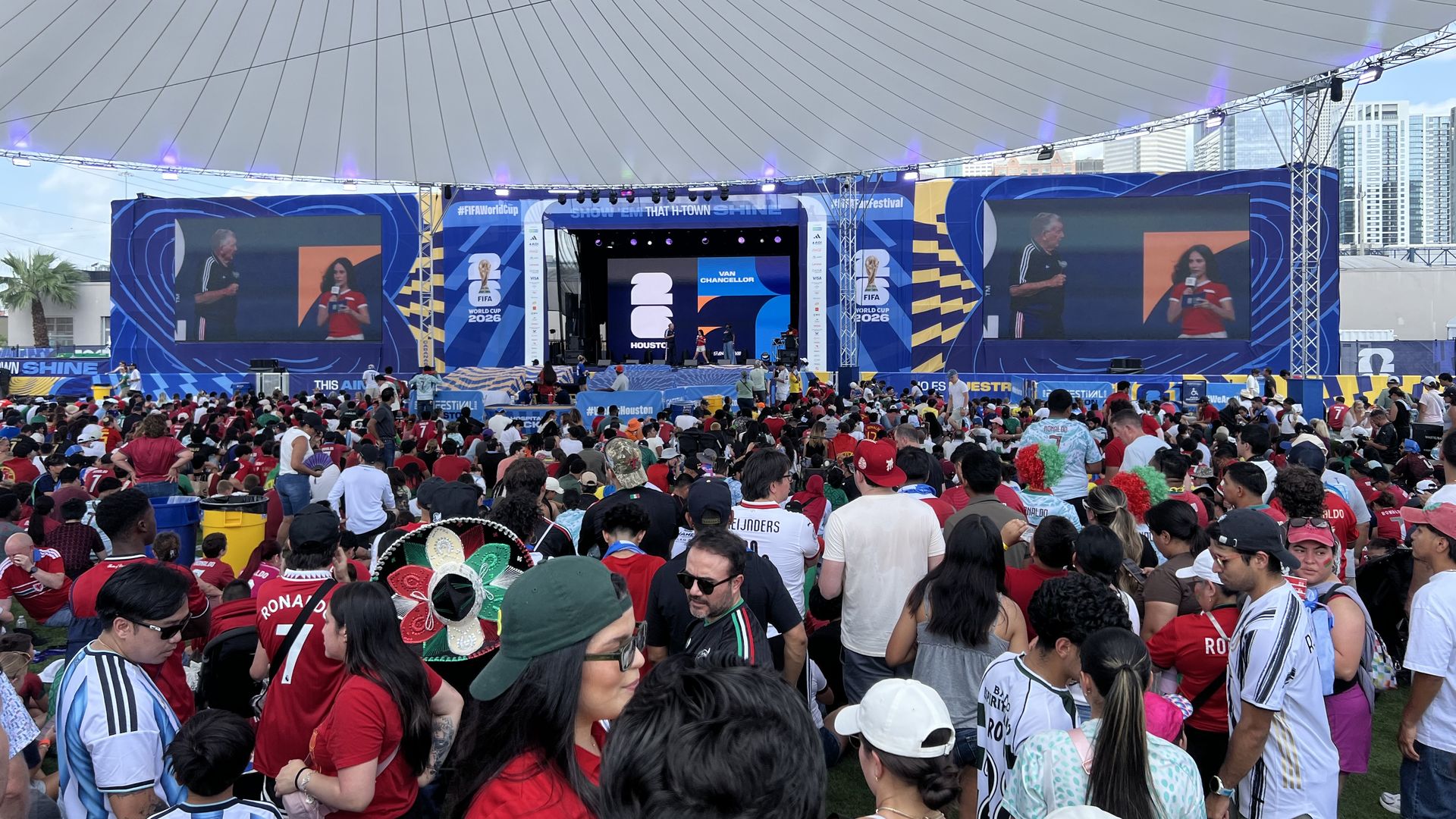 Outdoor stadium stage with blue banners and two large screens, performers on stage; crowd mostly in red shirts; a cable-supported dome overhead and city buildings in the background.