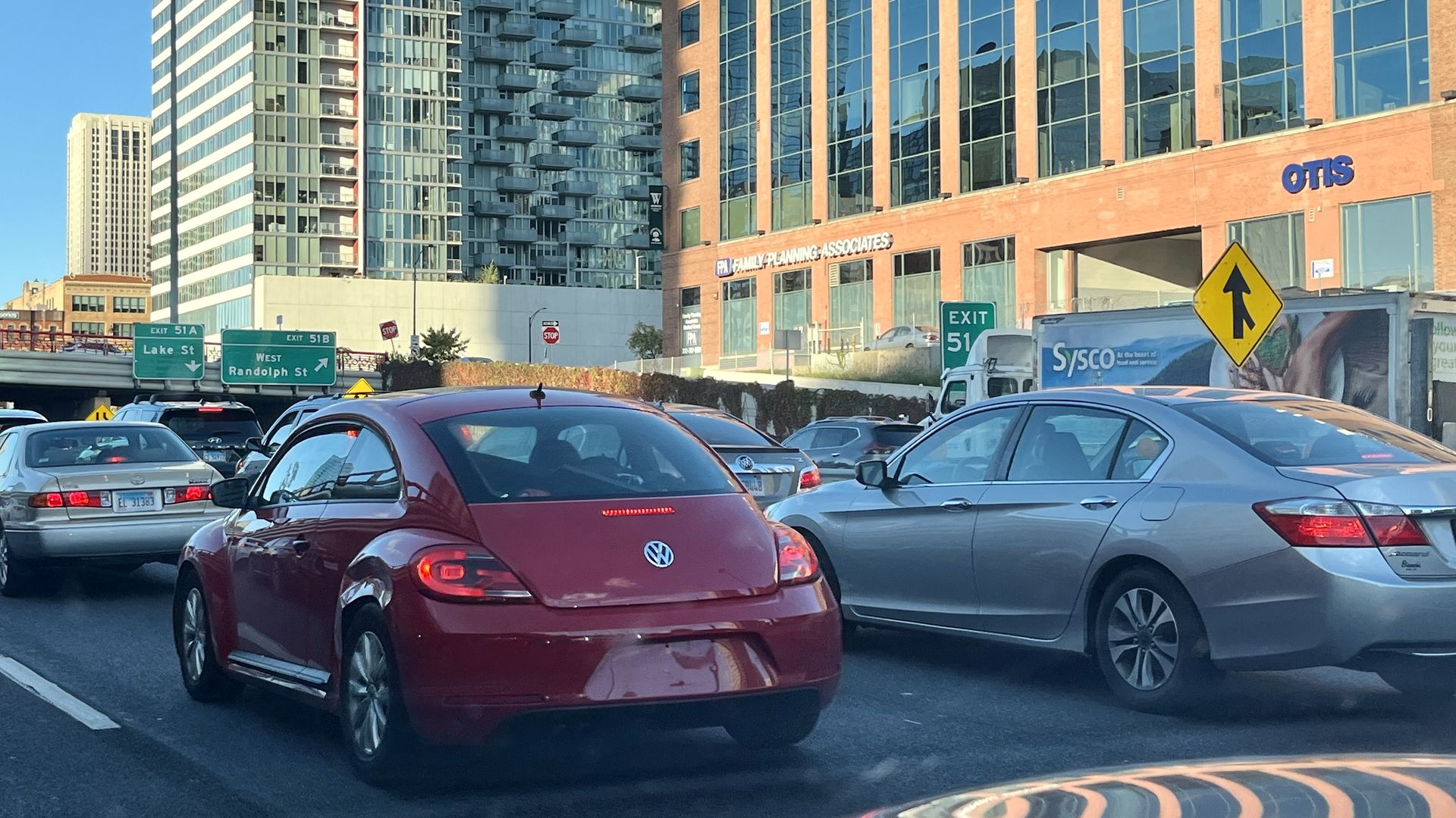 Traffic jam with various cars including a red Volkswagen Beetle on a city street near a building with signs "Family Planning Associates" and "OTIS" under clear blue sky.