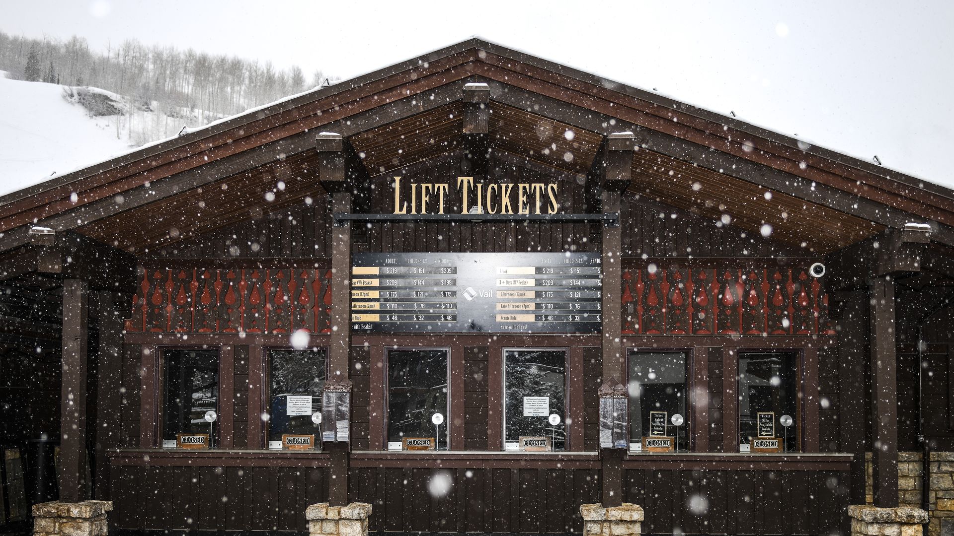 Snowy scene of a brown wooden lift-ticket booth with a peaked roof and a gold lift-tickets sign above a menu board; CLOSED signs on doors, with snow-covered hills and trees in the background.
