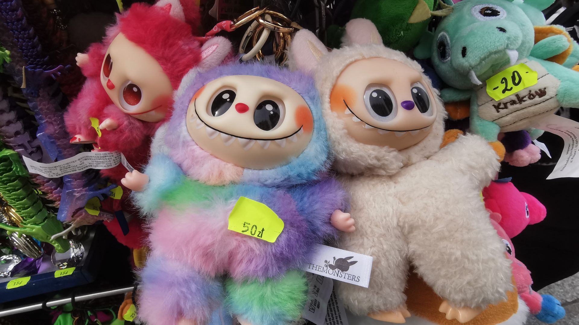 Colorful plush monster dolls with big eyes and sharp teeth, including a rainbow one marked 50 zł and a cream one, displayed among other toys with price tags in a market setting.