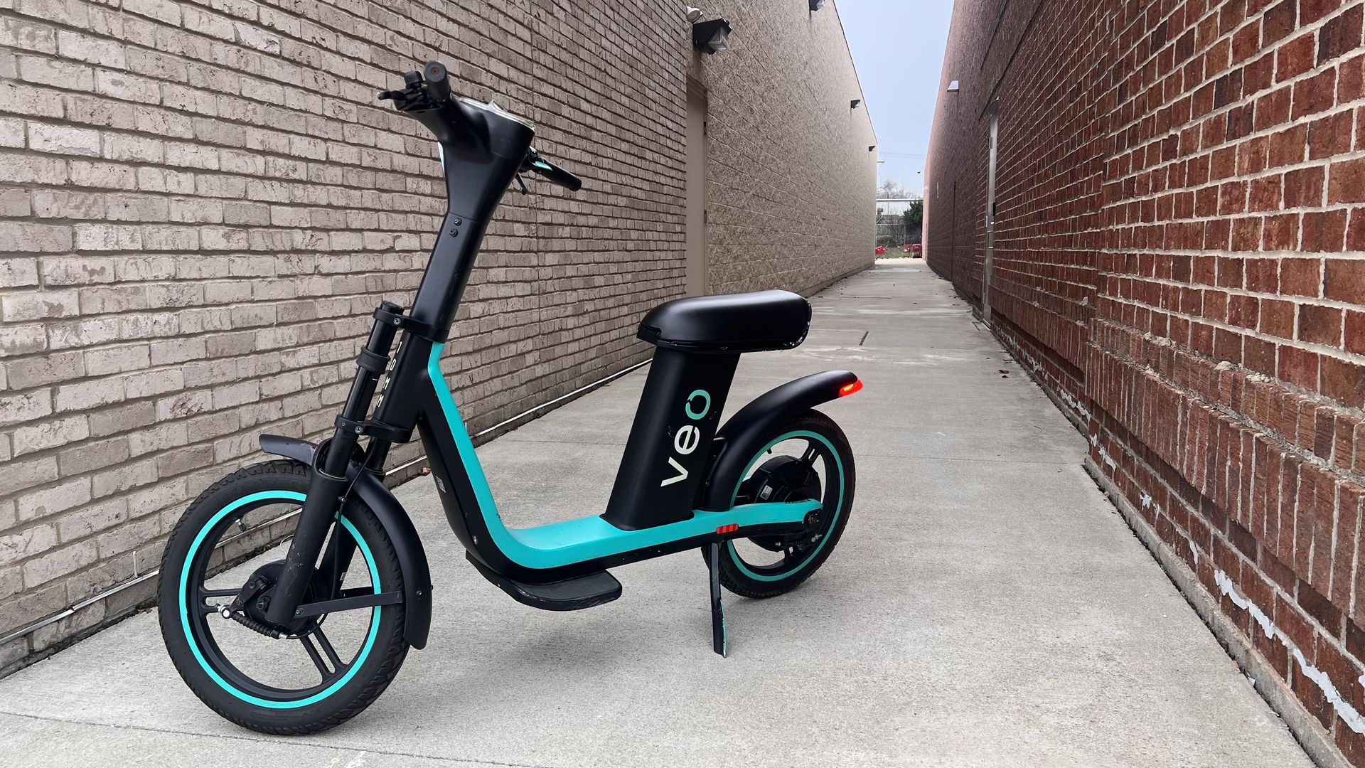 Black and teal electric bike with "veo" on the side, parked on a concrete alley between beige and red brick walls under a clear sky.